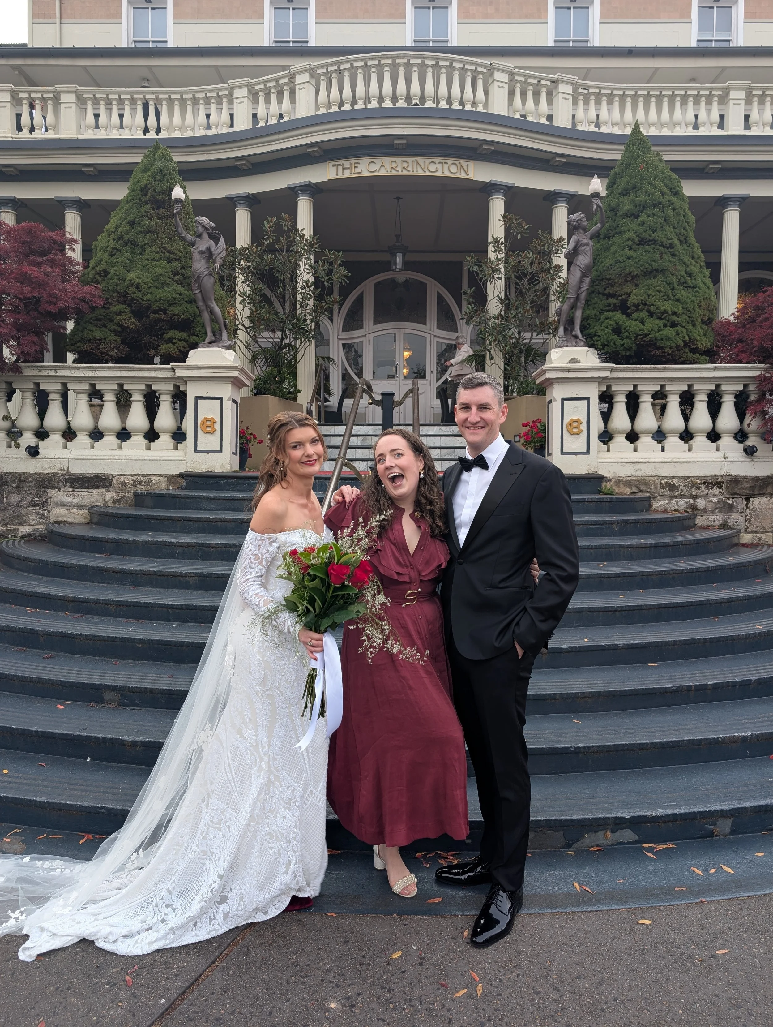 Elle is standing between a bride and groom in front of a large staircase to the art deco style Carrington Hotel in Katoomba, and they are all smiling and laughing together after their Blue Mountains marriage ceremony