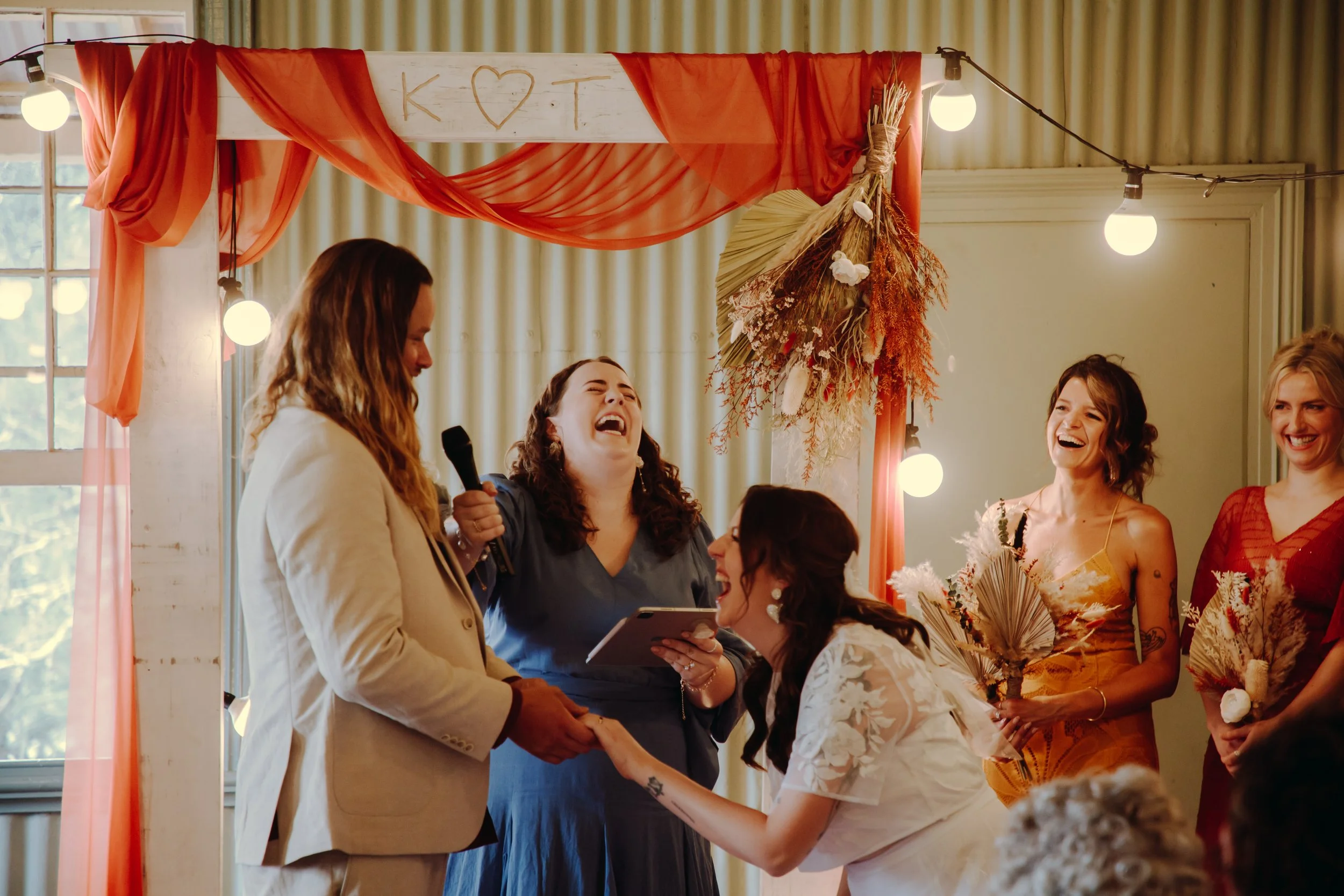 A wedding ceremony with a bride in a white dress, groom in a beige suit, Elle the celebrant in a blue dress holding the microphone. There is a white arch with orange draping, and bridesmaids in the background in sunset colours. Everyone is laughing
