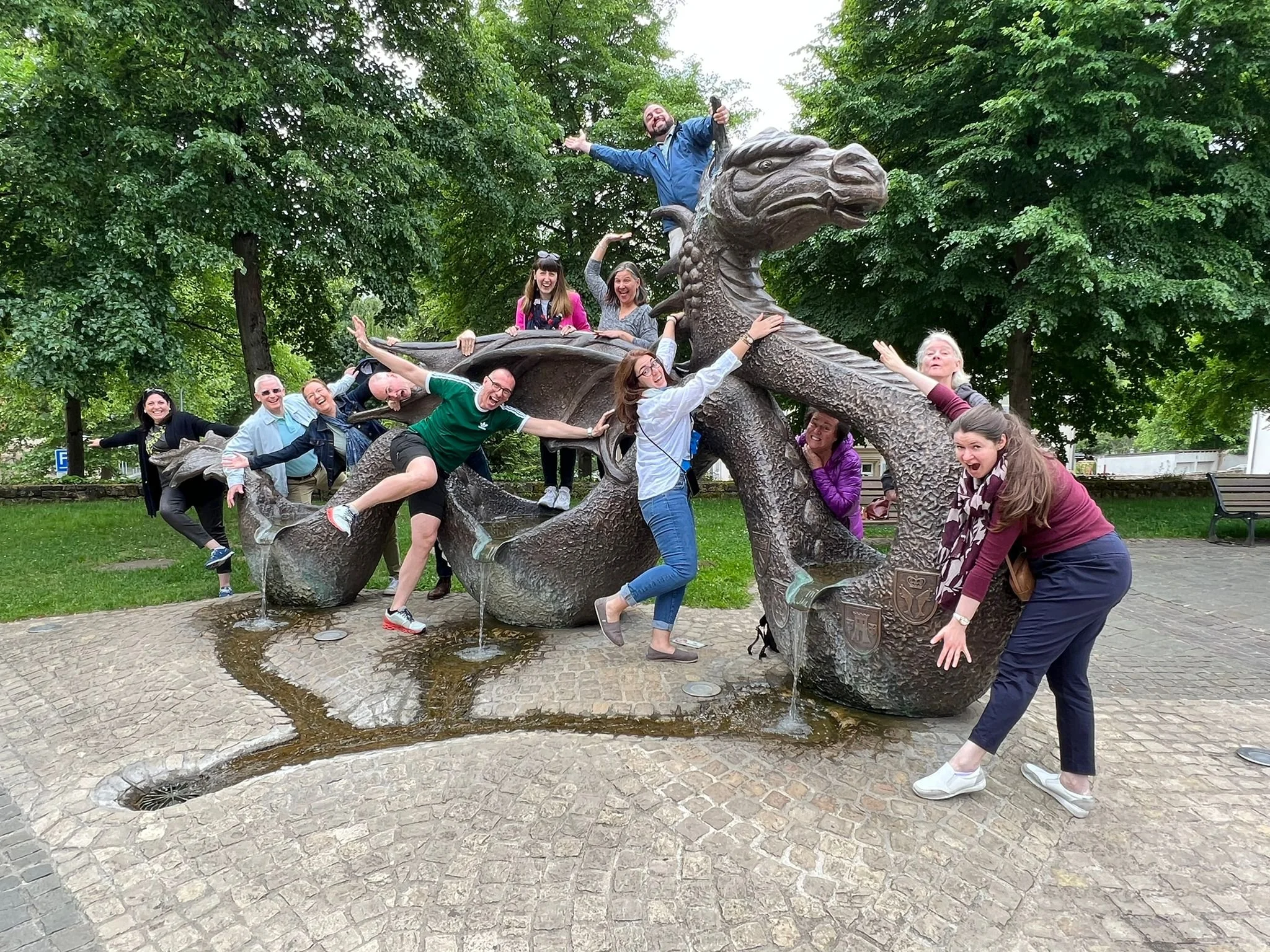 A group of people playfully interacting with a large bronze dragon fountain in a park, with some missing water from its mouth and body.