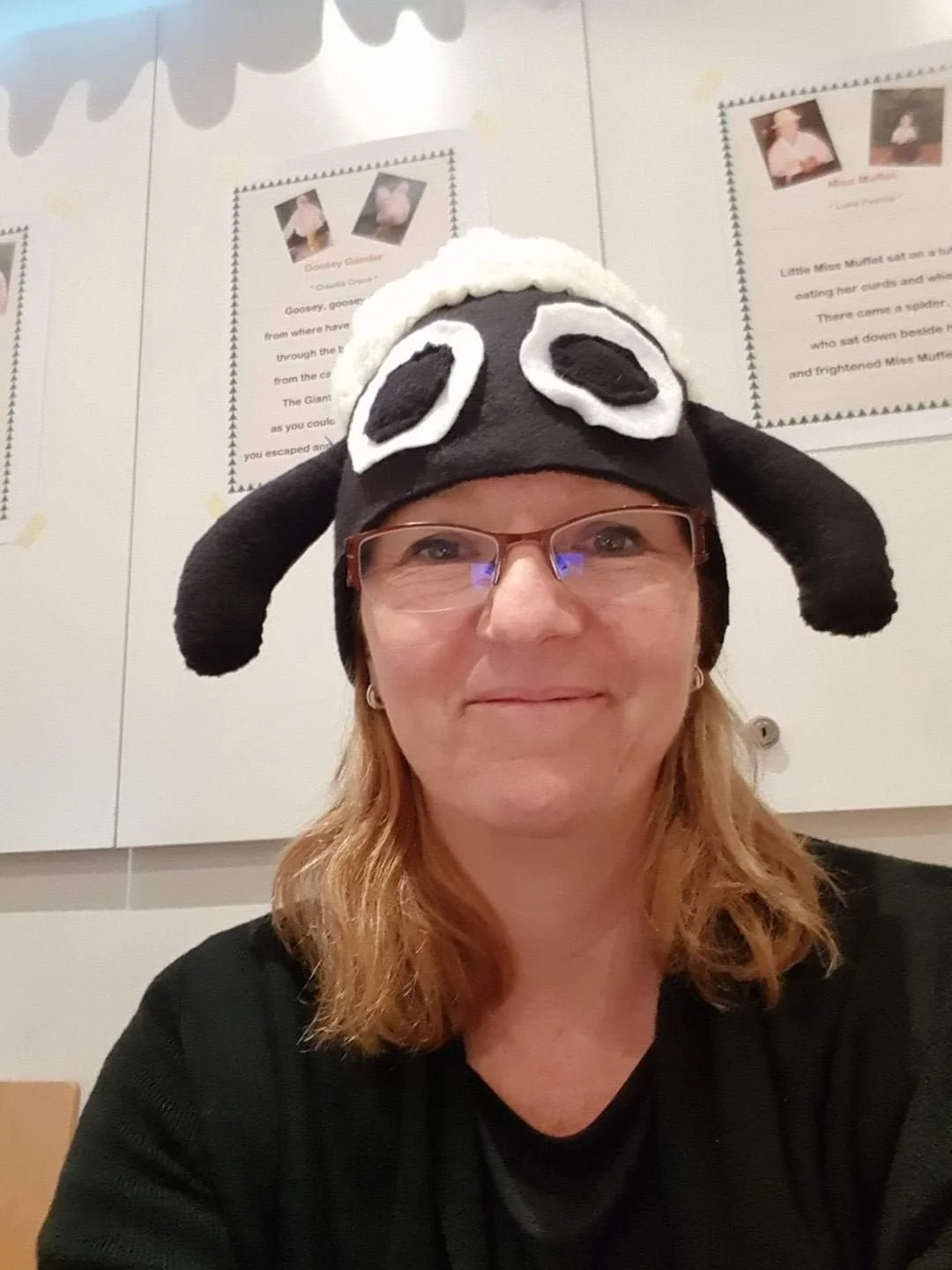 Woman wearing a black and white sheep hat with floppy black ears, glasses, and a black shirt, sitting in front of posters with text.