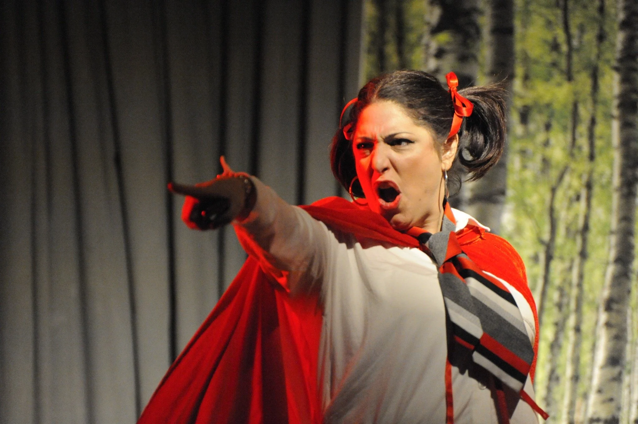 A woman with dark hair styled in pigtails tied with red ribbons, wearing a white shirt, red tie, and red cape, appears to be shouting and pointing aggressively during a theatrical performance with a forest backdrop.