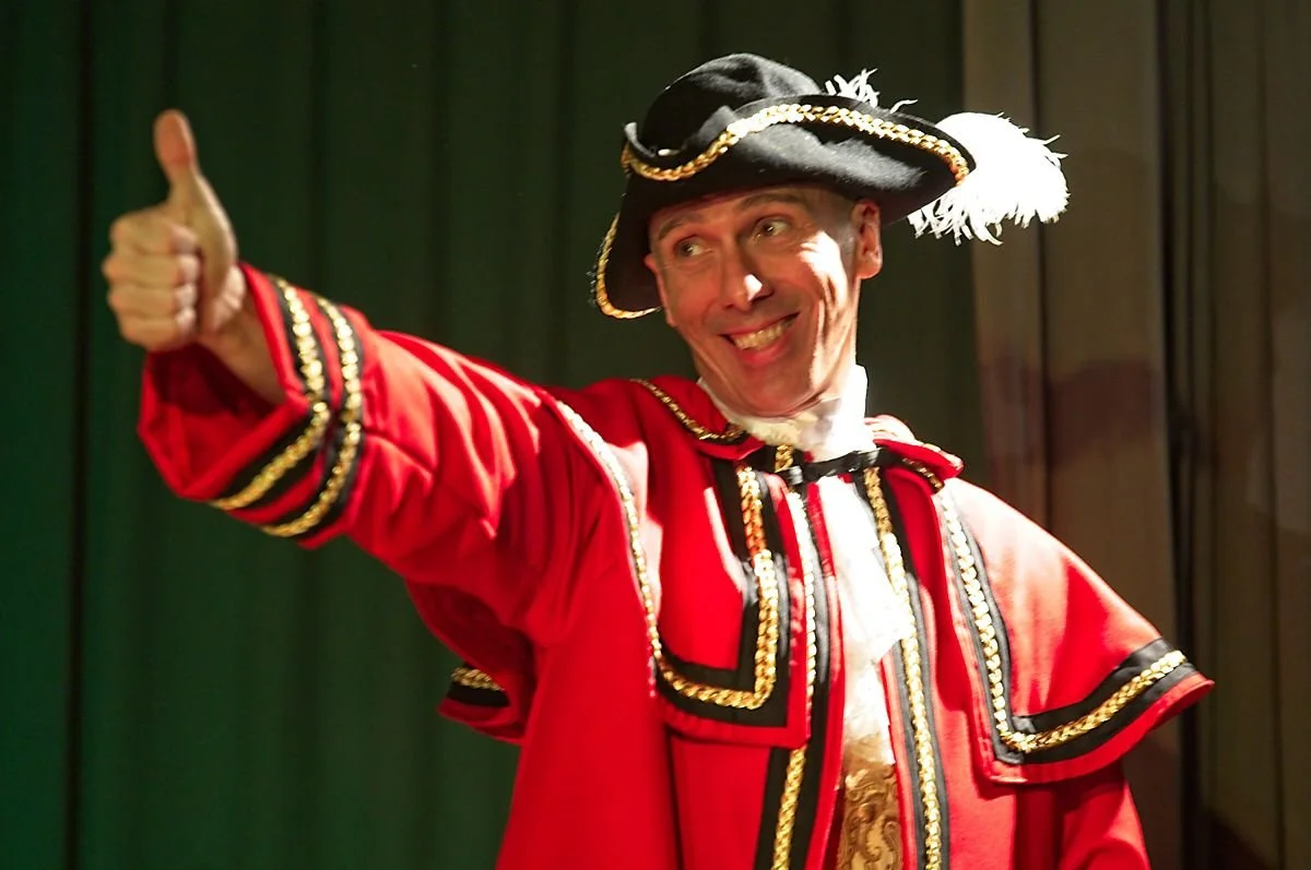 A man dressed as a town crier in a red and black costume with gold accents, wearing a feathered hat, giving a thumbs-up and smiling.