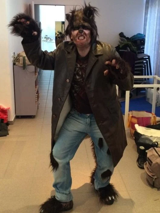 Person dressed in a full werewolf costume with furry paws, mask, and costume details, standing indoors and making a fierce gesture.