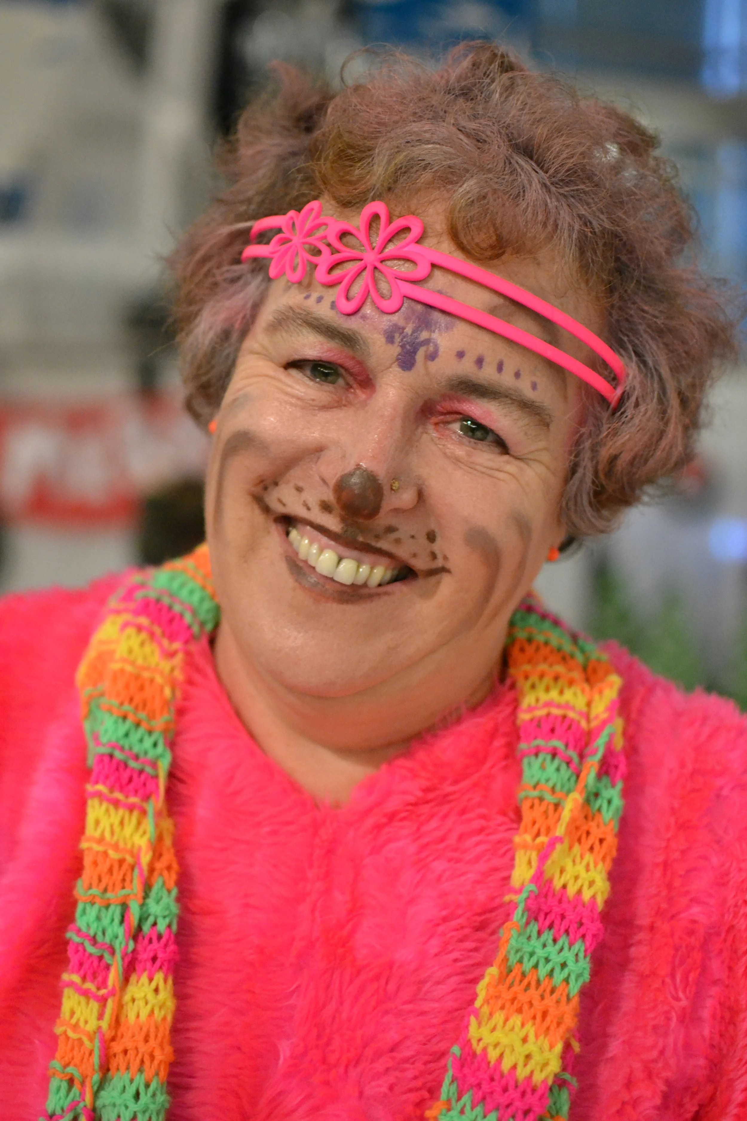 Woman smiling with colorful face paint, pink headband with flower design, and a multicolored knitted necklace.