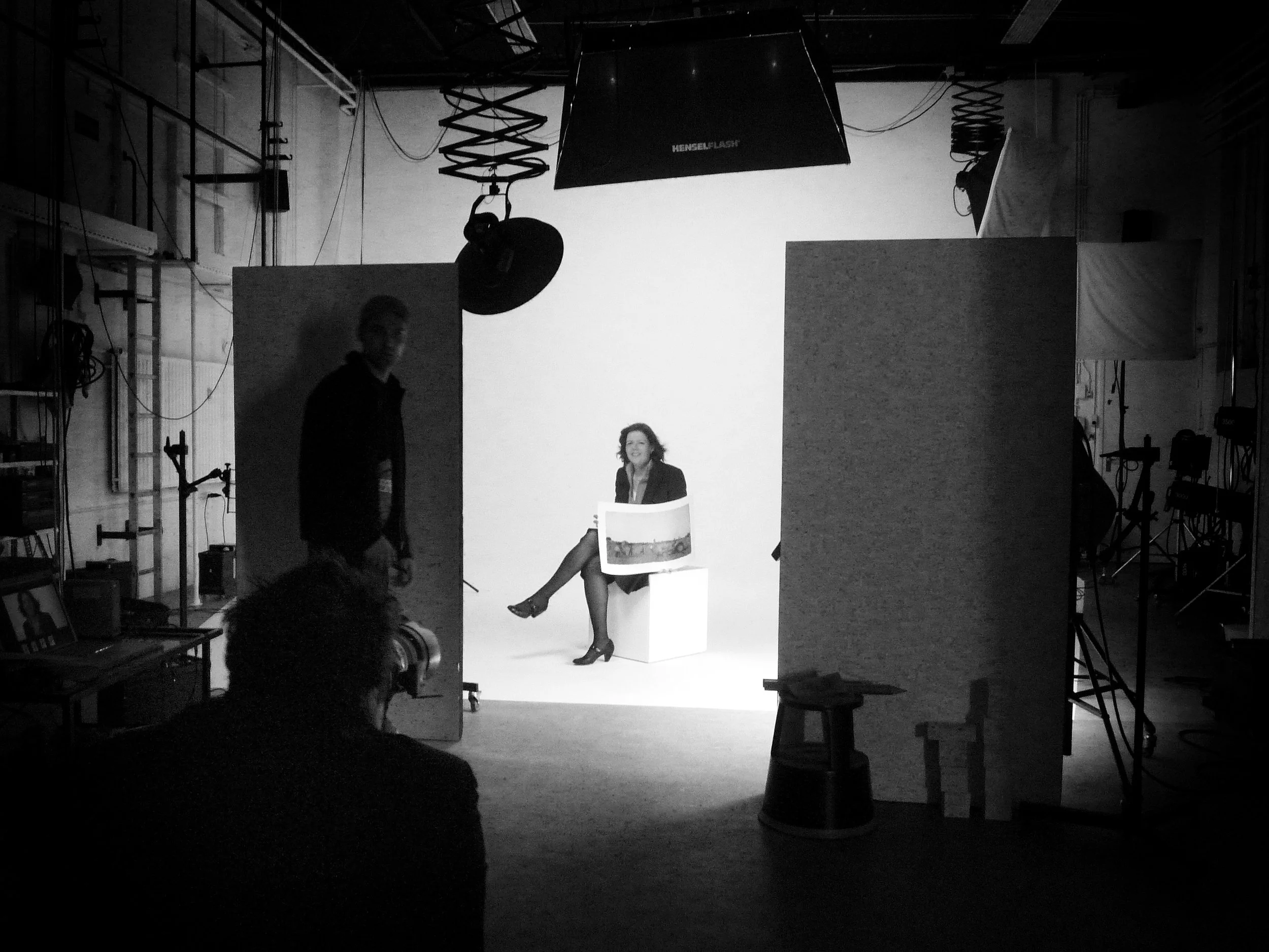 In een zwarte en witte studio, een vrouw zit lachend op een witte blok, met een schilderij op haar schoot, terwijl haar benen over elkaar hangen. Twee ronde wanden scheiden haar van de fotograaf en een man die op de linkerzijde staat. Er zijn profess