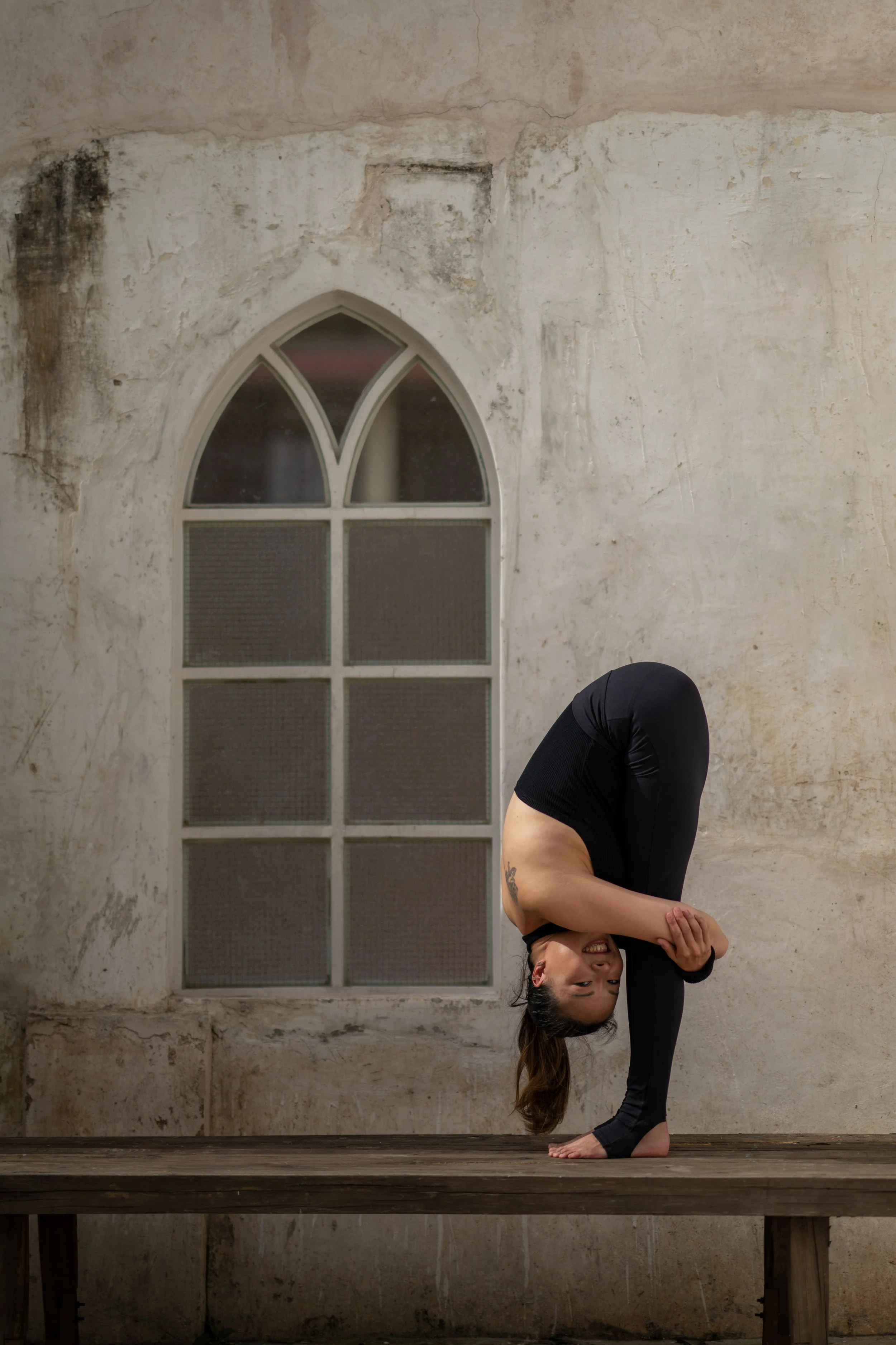 Woman practicing yoga in a stretch pose on a wooden platform in front of an old, weathered wall with a stained glass window.