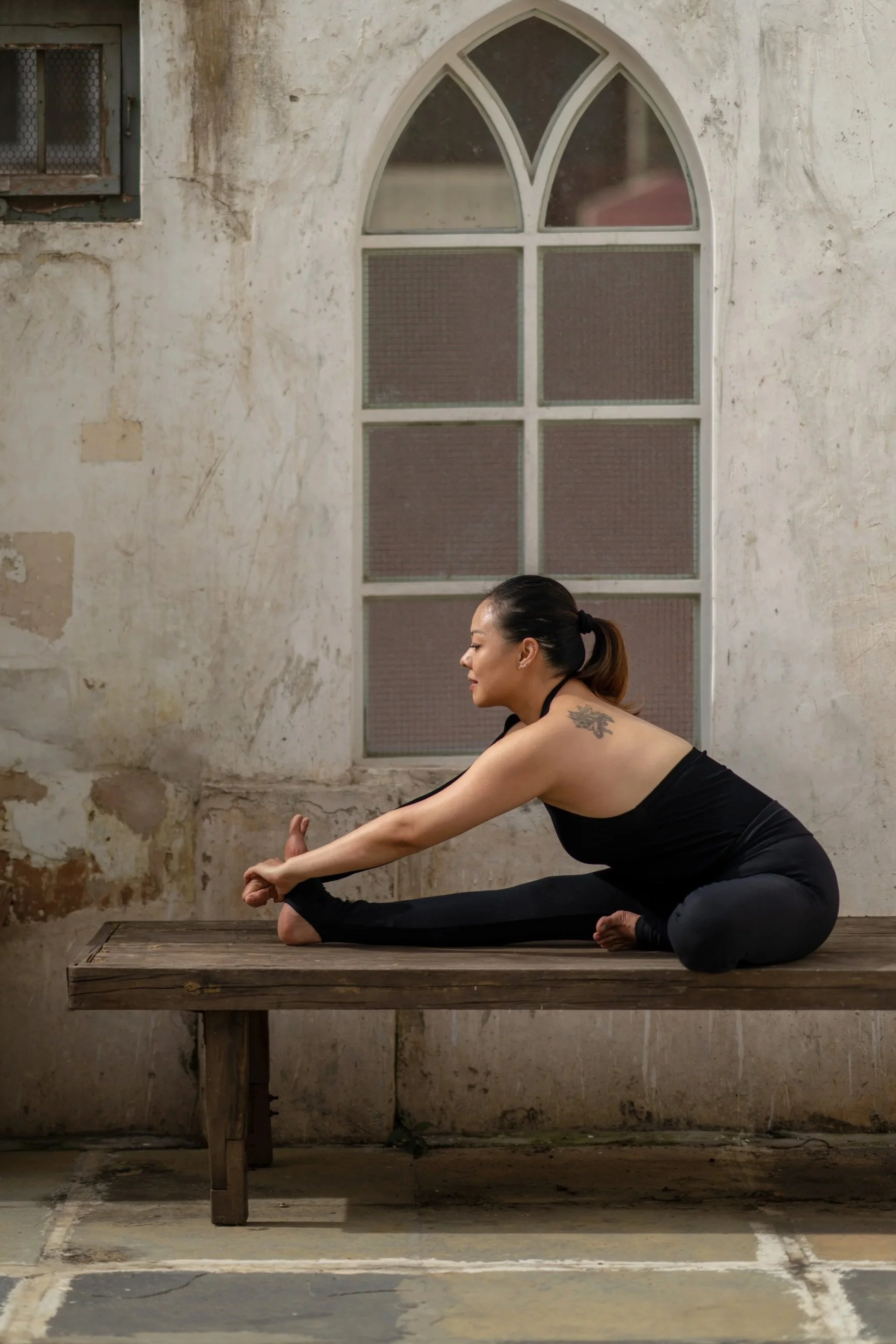 Woman practicing yoga indoors, sitting on a wooden platform, stretching her leg while reaching forward with her hands, wearing black athletic clothing, with a tattoo on her shoulder, in front of an old wall with a large arched window.