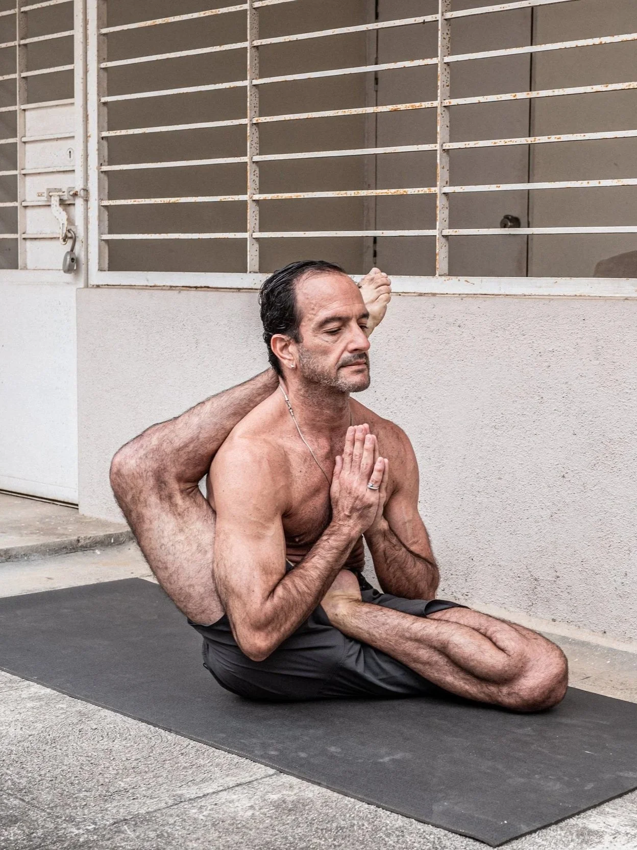 Two shirtless men practicing partner yoga outdoors, with one man sitting cross-legged with hands in prayer position, and the other man leaning on his back with arms extended behind his head, against a neutral background.