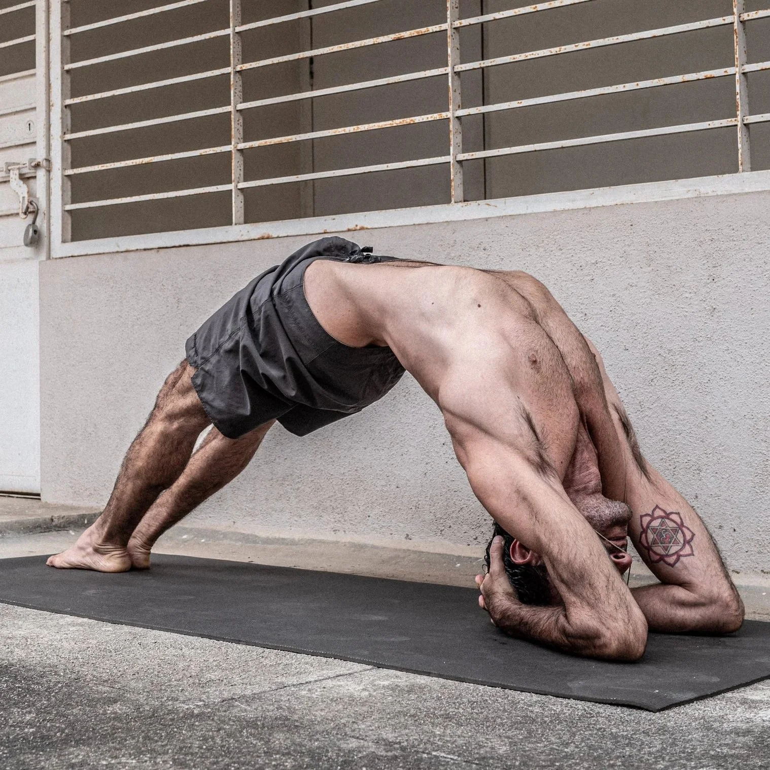 A shirtless man with a tattoo on his arm performing a downward dog yoga pose on a black mat outside near a wall with a vent and grate.