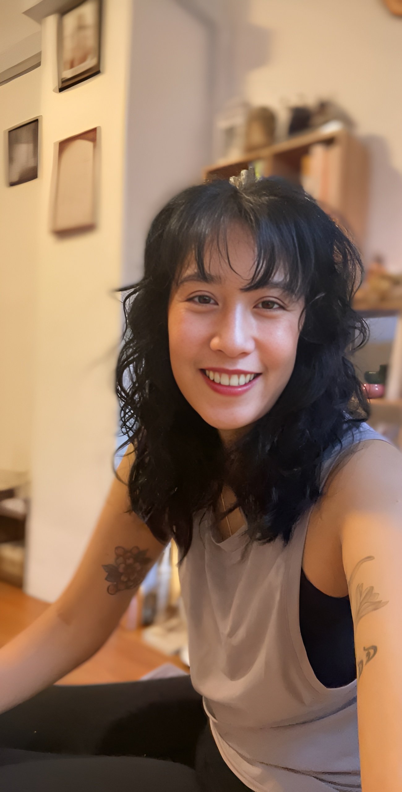 A smiling woman with wavy black hair, tattoos on her arms, wearing a sleeveless top, seated indoors with a blurred background of books and picture frames.