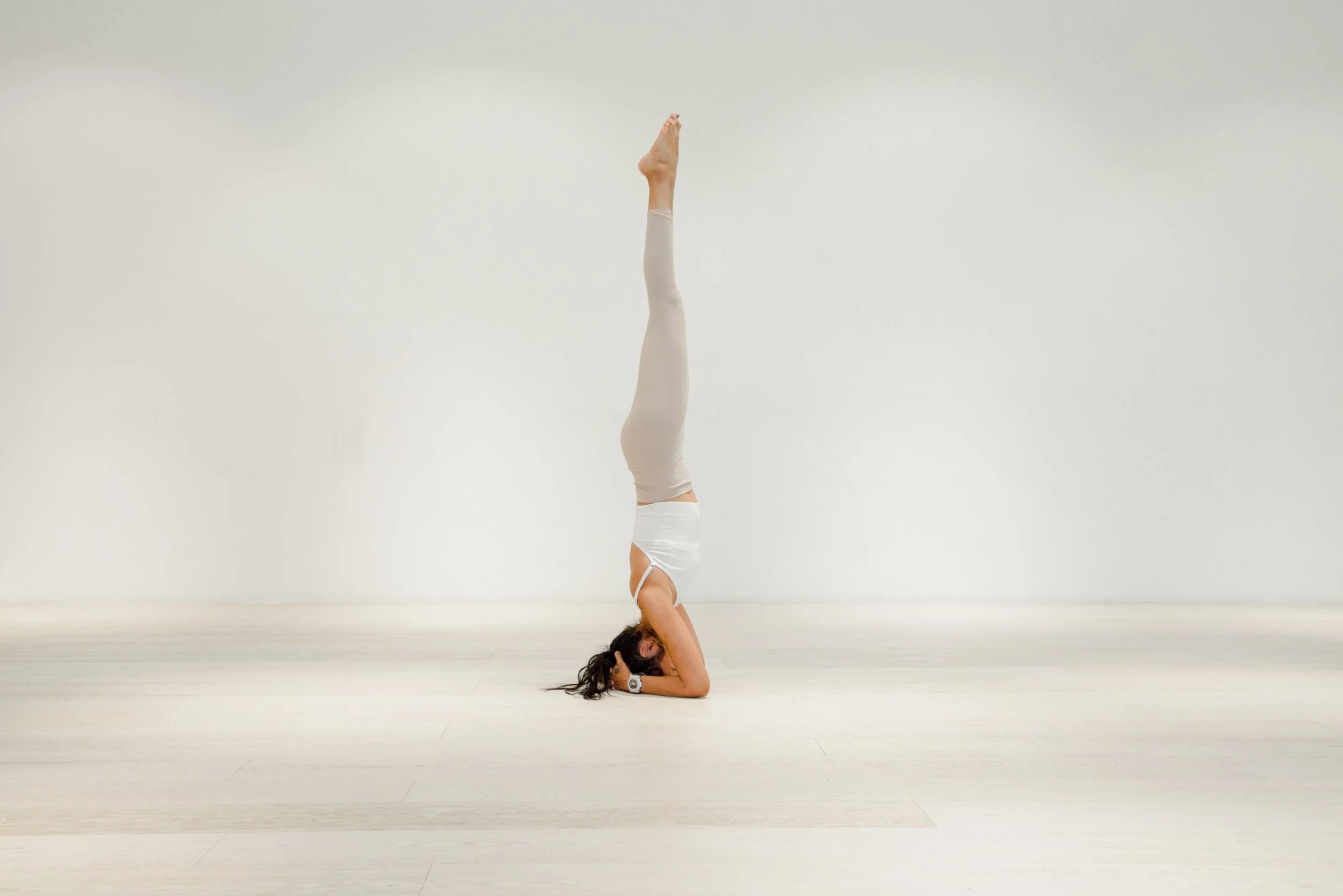 A woman practicing yoga in a headstand pose in a bright, minimalistic room with light-colored wooden floor and plain white wall.