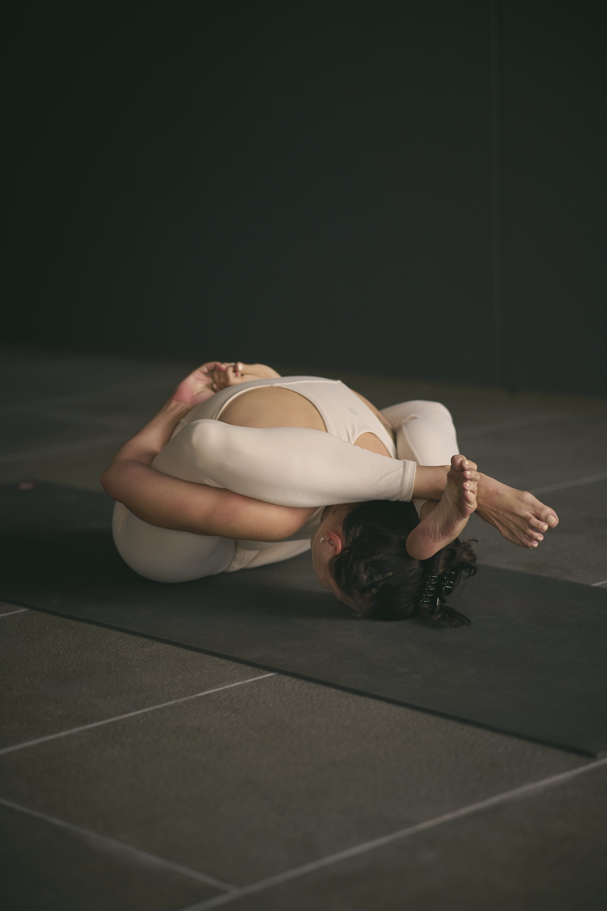 A woman practicing yoga in a kneeling forward bend pose on a black mat against a dark wall.