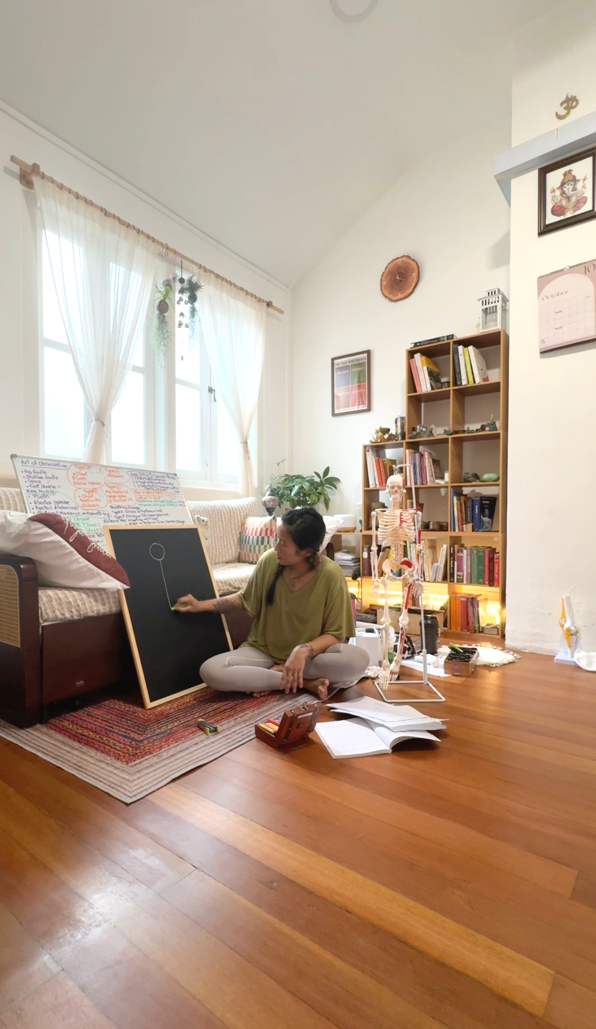 A woman sitting cross-legged on a rug beside a blackboard, teaching or explaining something with a diagram, inside a cozy living room with bookshelves, a human skeleton model, and natural light from windows.