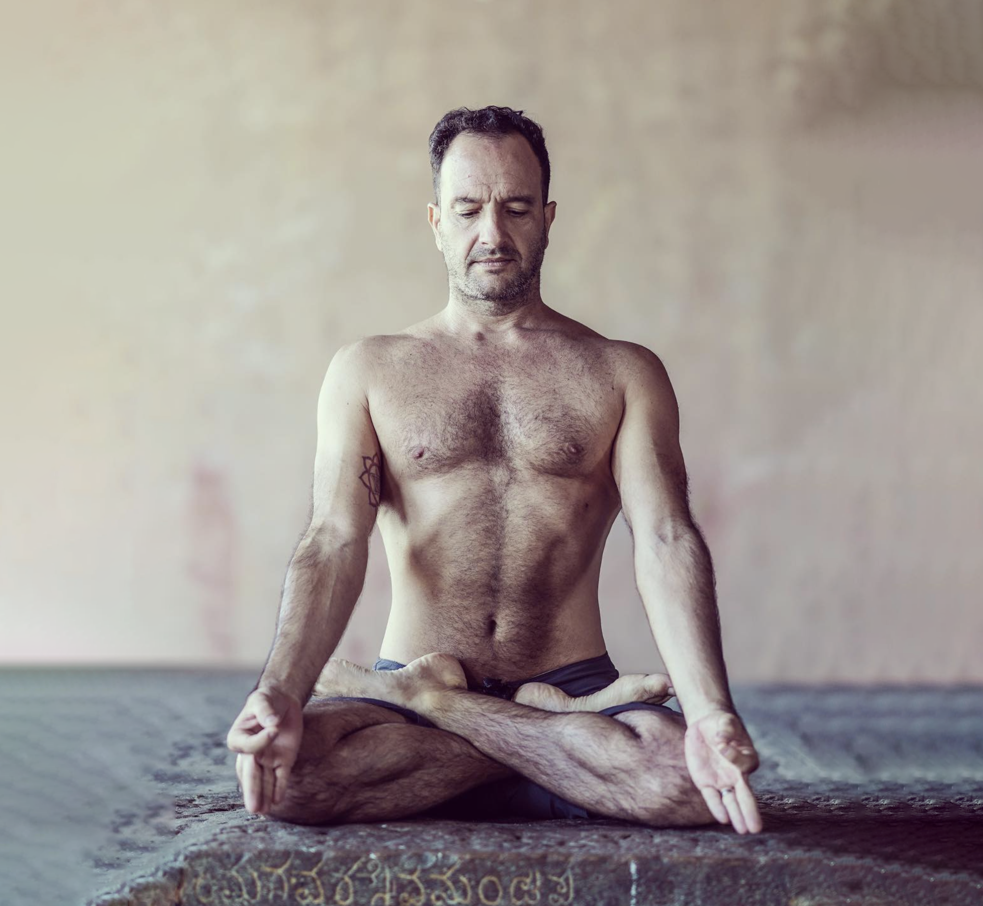 A man with a beard and short hair practicing yoga, sitting cross-legged on a mat in a meditation pose with hands resting on knees, in a minimal interior space.