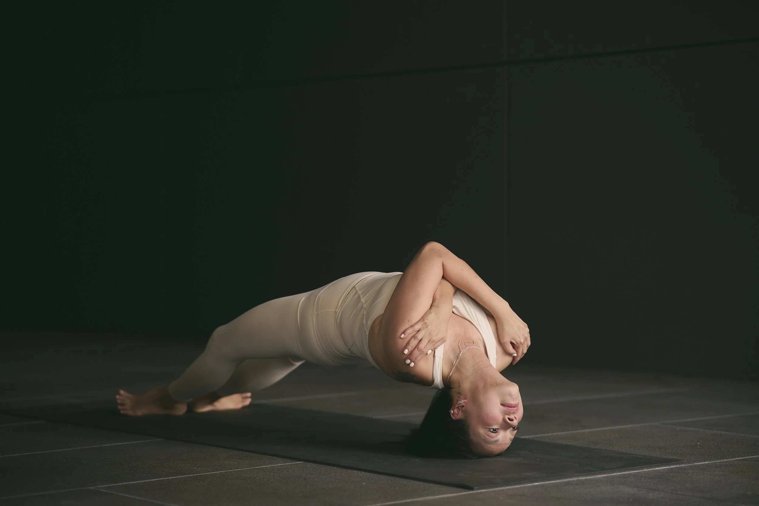 A woman practicing yoga on a black mat in a dark room, performing a side plank pose with her arm wrapped around her torso and her head resting on the floor.