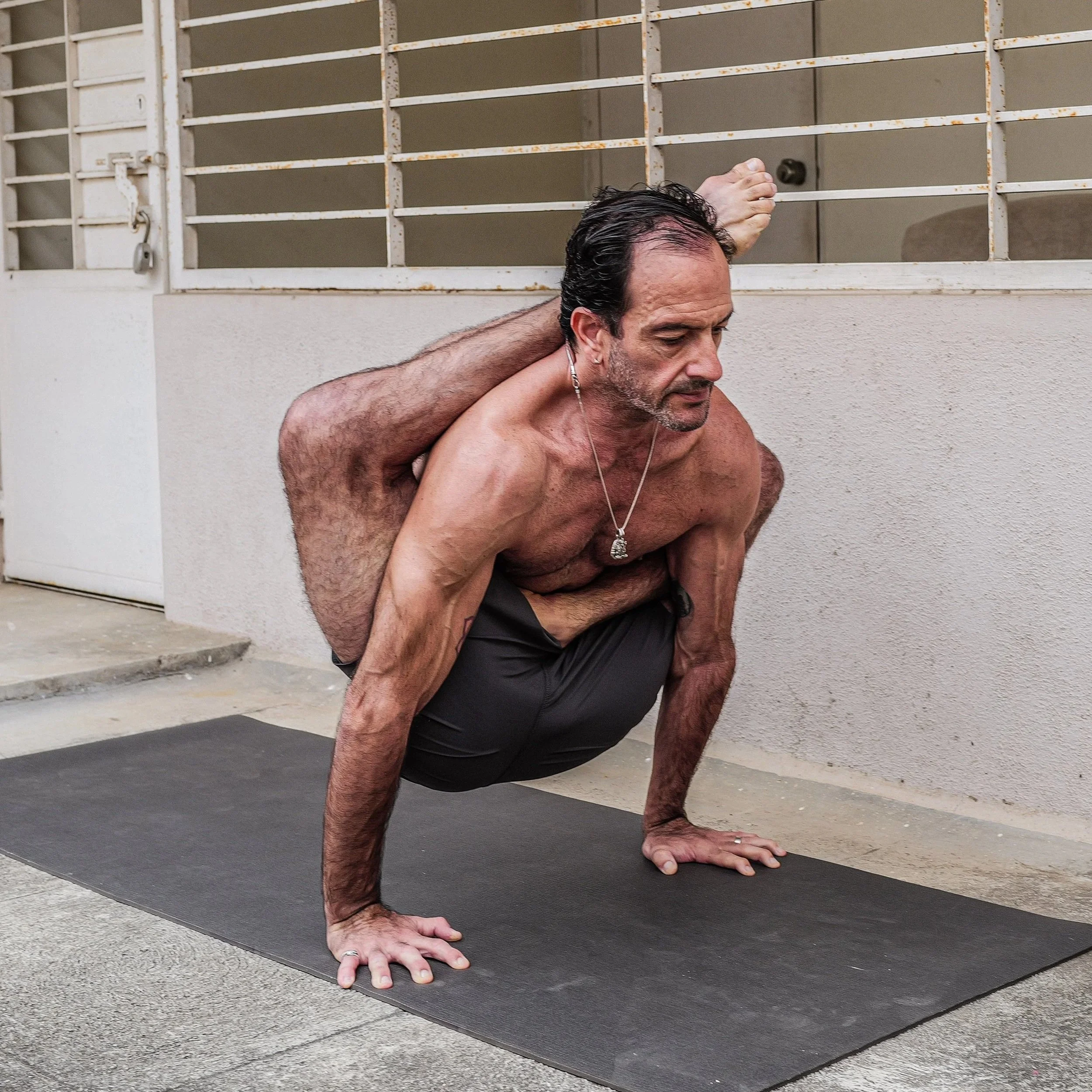 A shirtless man with a necklace doing a challenging yoga pose outside on a black mat, balancing on his hands with one leg bent and foot resting on the back of his head.