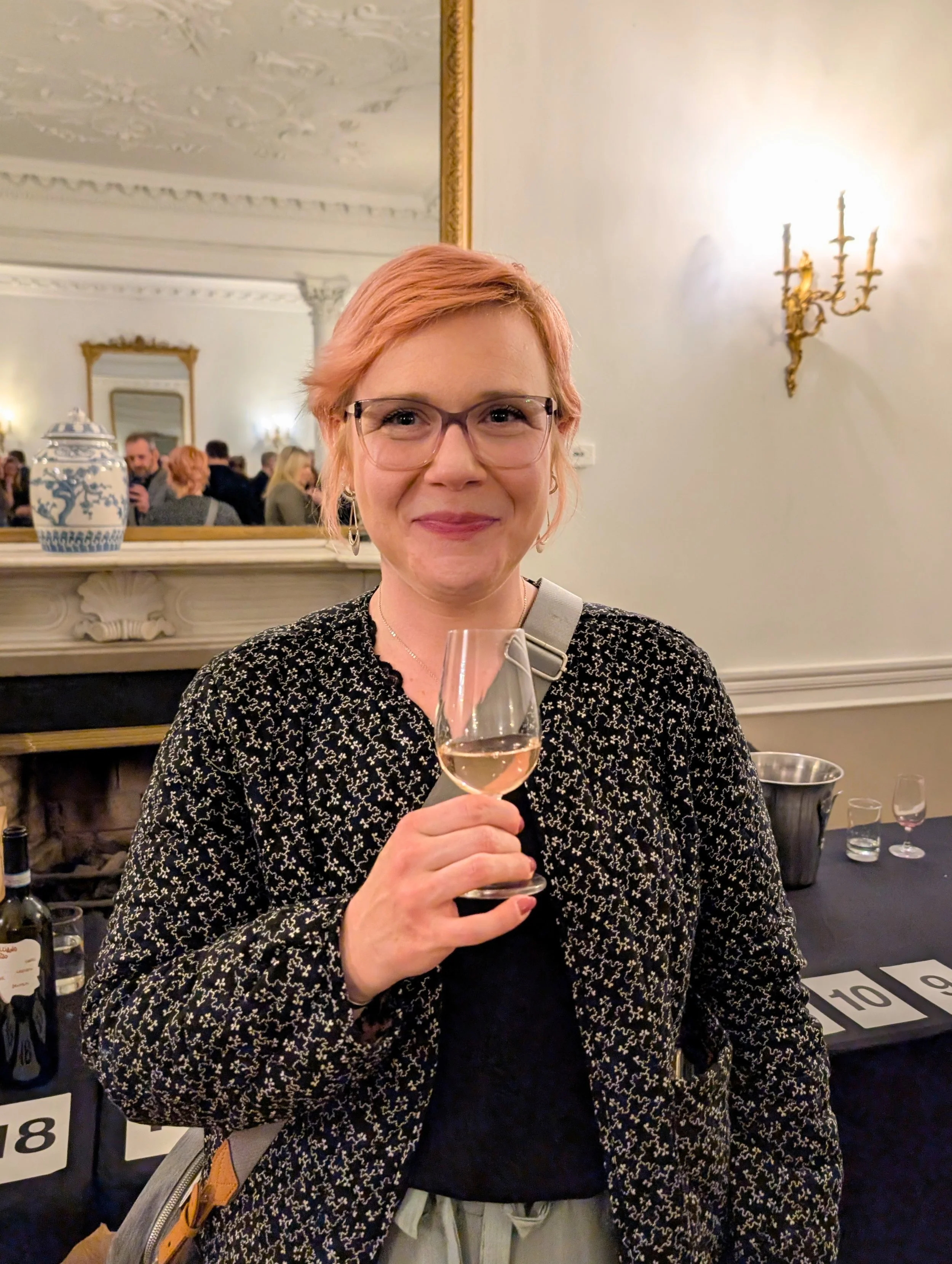 A woman with short pink hair and glasses holding a glass of rosé wine at a social gathering in an elegant room with ornate ceiling molding and a gold-framed mirror.