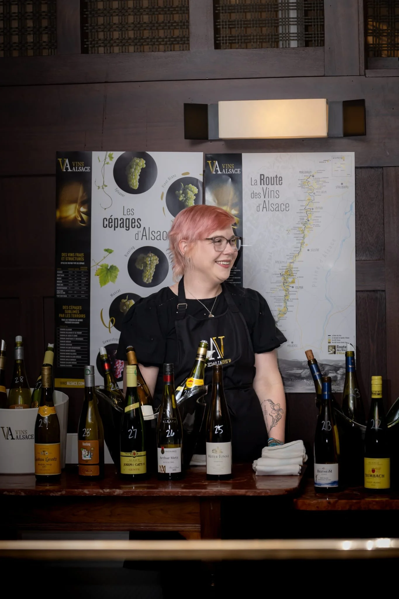 A woman with pink hair, glasses, and tattoos smiling behind a wine tasting table with bottles of wine numbered 21 through 28, and wine posters and a map of Alsace, France in the background.
