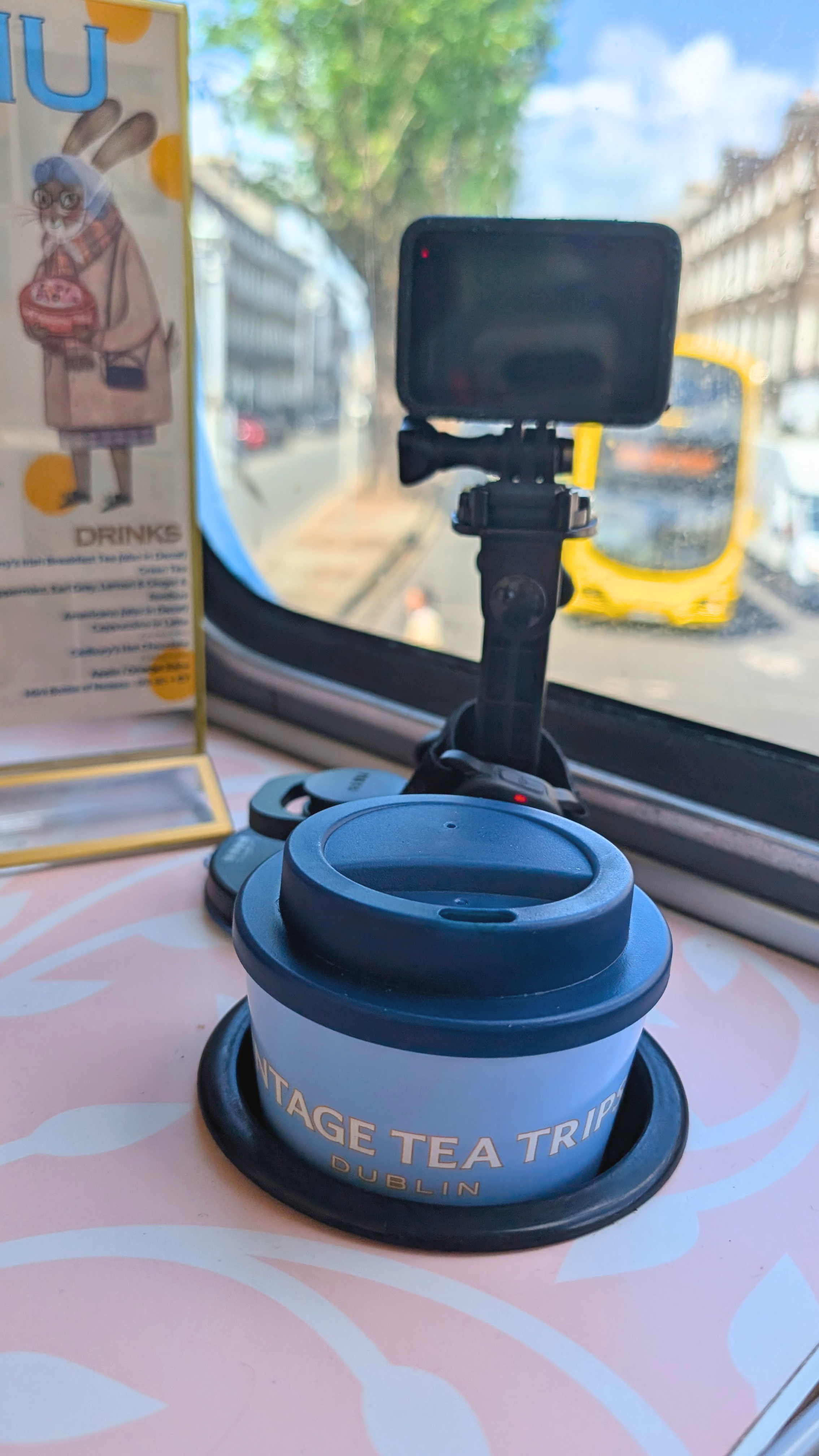 GoPro camera mounted to a table with retro pink and white print, pointing out the window at a Dublin city bus. There is a drinks menu with an illustrated rabbit character wearing glasses in a gold frame, and a to go cup that says 'Vintage Tea Trips'