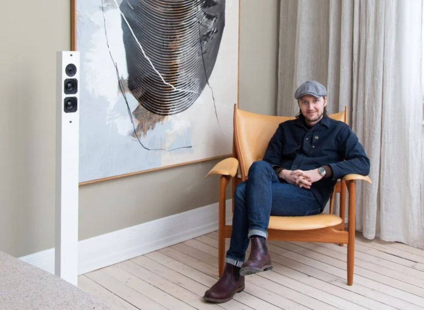 A man sitting in a wooden chair with beige upholstery, wearing a gray cap, black shirt, jeans, and brown boots. He is smiling and sitting with legs crossed in front of a large abstract artwork and a white speaker, in a room with light-colored curtains.