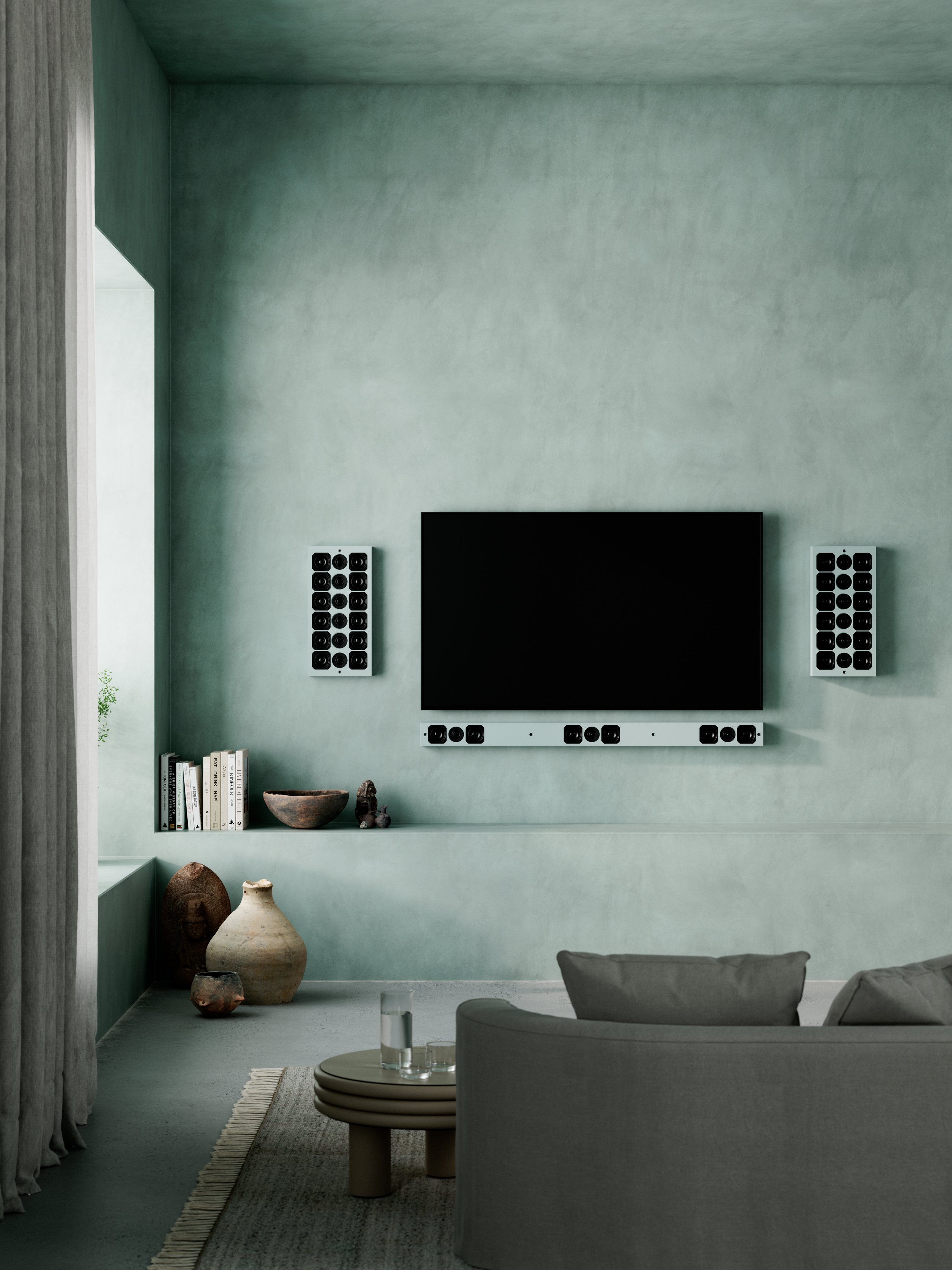 Modern living room with a mounted flat-screen TV, two black and white speakers on each side, decorative vases, books, and a remote control on a small table, with a beige sofa and a window with curtains.