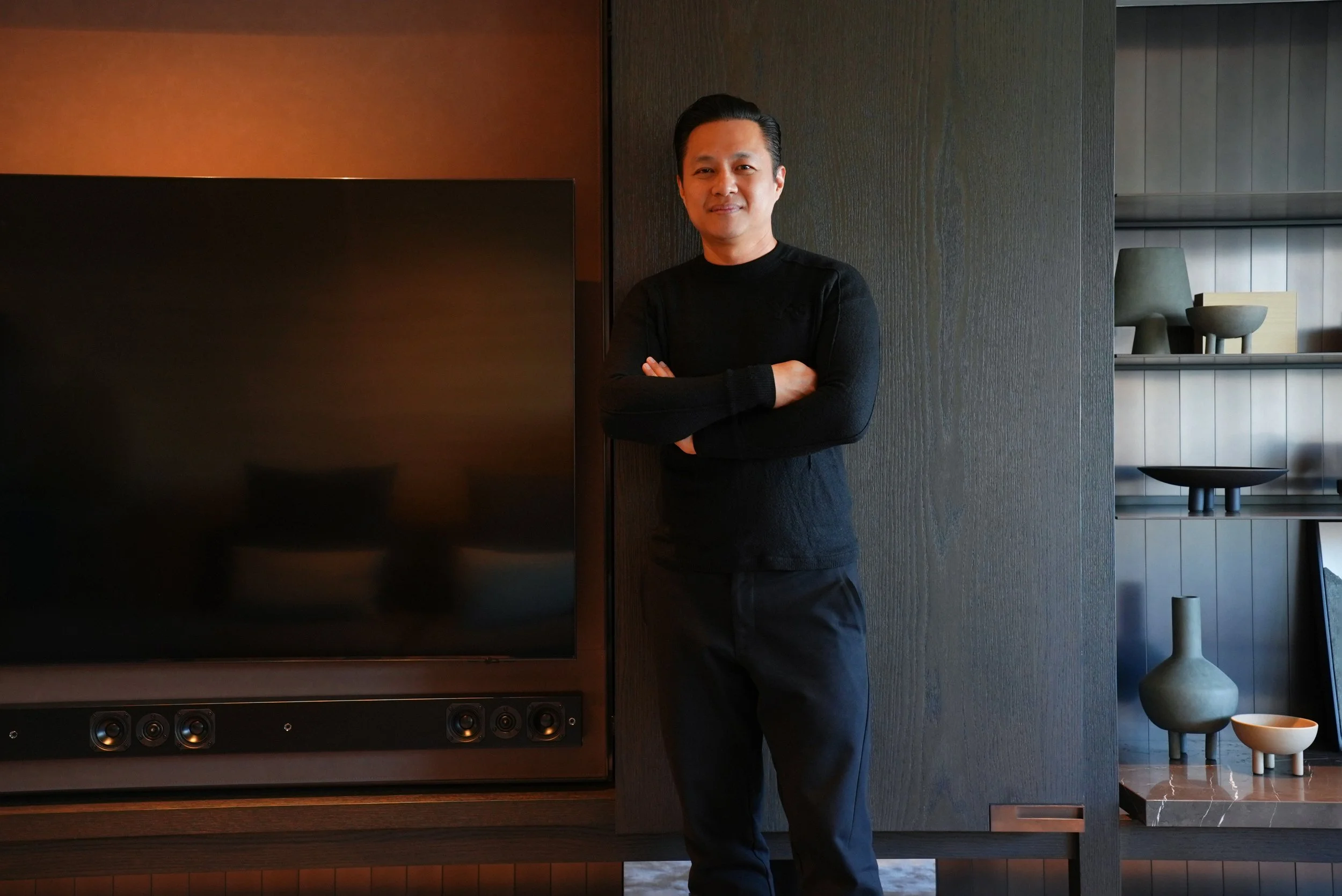 A man stands with arms crossed next to a large flat-screen TV in a modern room with decorative shelving.