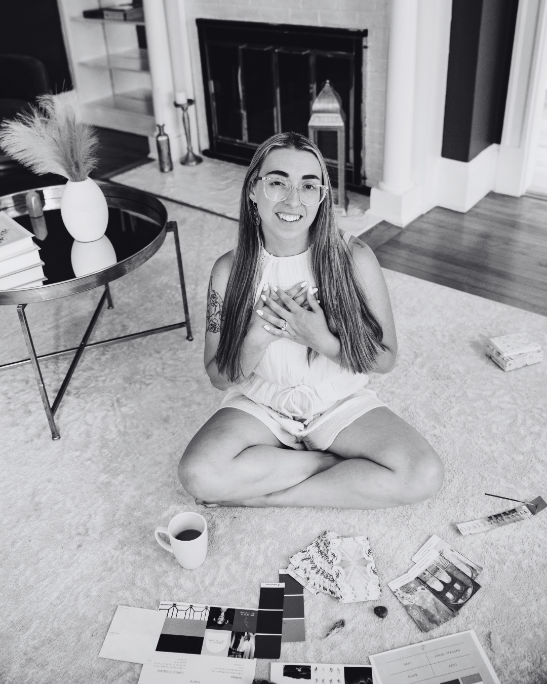 A woman sitting cross-legged on the carpeted floor with her hands on her chest, smiling at the camera. She is surrounded by various items including a mug of coffee, color sample swatches, brochures, and a small decorative object in a cozy living room setting with a fireplace, books, and a vase with pampas grass.