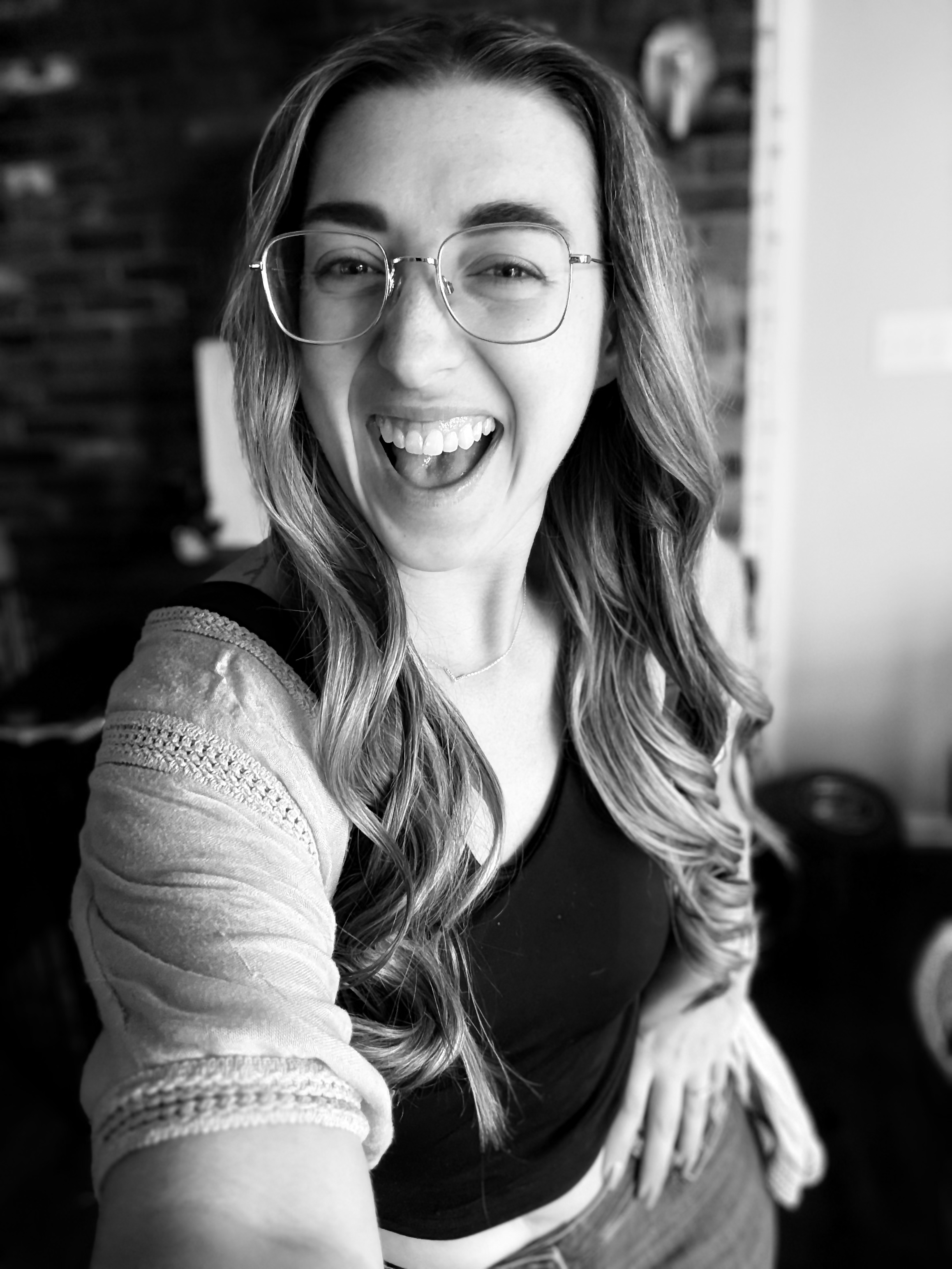 A woman with long wavy hair, wearing glasses, smiling with her mouth open, taking a selfie. She has a light-colored cardigan over a dark top, and a necklace. The background includes a brick wall and some blurred objects.