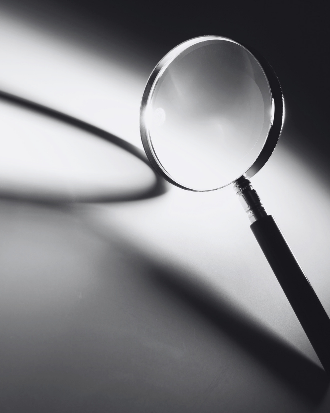 A magnifying glass with a black handle, casting a shadow on a smooth surface with light and dark areas.