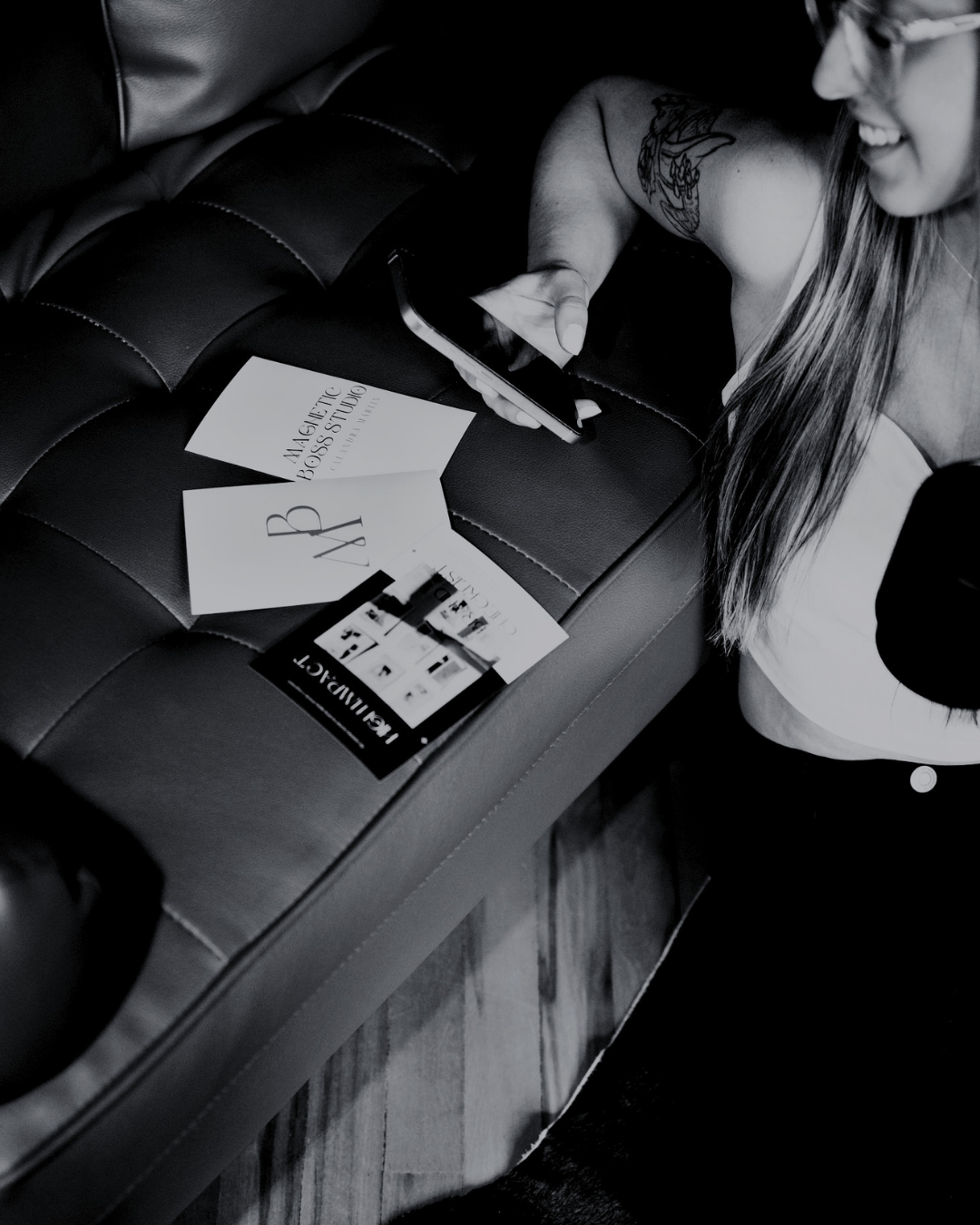 A woman with long hair and glasses smiling while looking at her phone, sitting at a table with promotional materials for the 'MAGNETIC BOSS STUDIO' and 'LAB M. D. S' magazine, on a leather bench seat.
