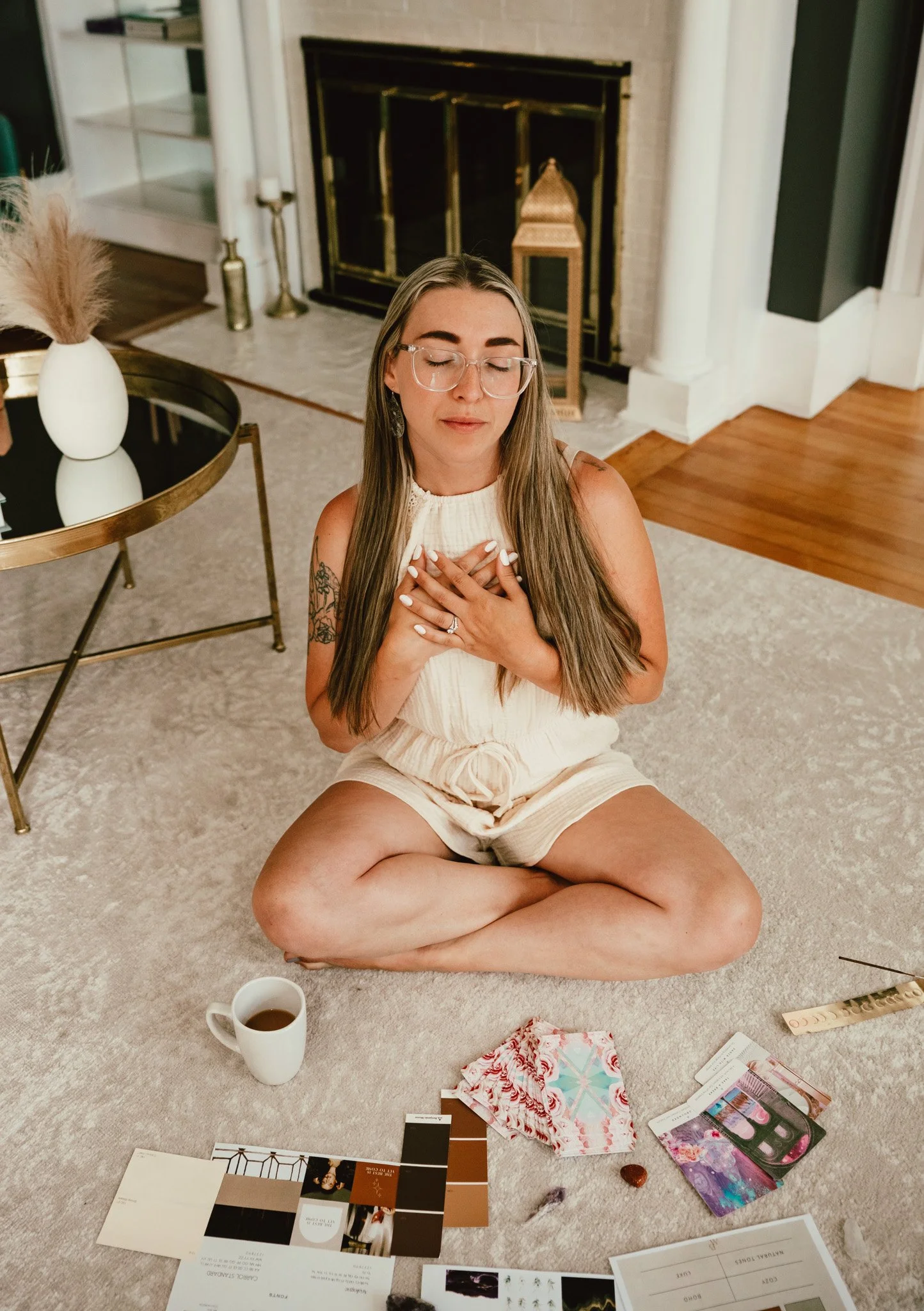 A woman with long hair and glasses sits cross-legged on the carpeted floor with her eyes closed, holding her hands over her chest. In front of her are paint swatches, color palettes, a sheet of paper with a color chart, and other art supplies. To her left, there is a coffee mug on the floor, and a table with decorative items including a vase with pampas grass and a candle. The background features a fireplace, a lantern, and a corner of a room.