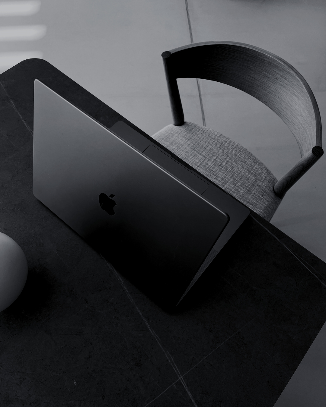 Top view of a closed black Apple MacBook on a dark table with a gray textured chair behind it.