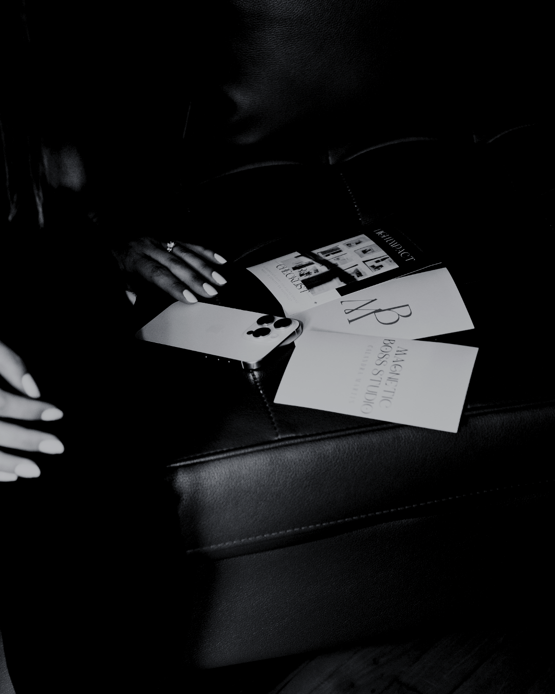 A person with manicured nails holding an iPhone with a multi-camera setup, sitting on a dark leather seat with Louboutin and BOSS STUDIOS brochures on their lap.