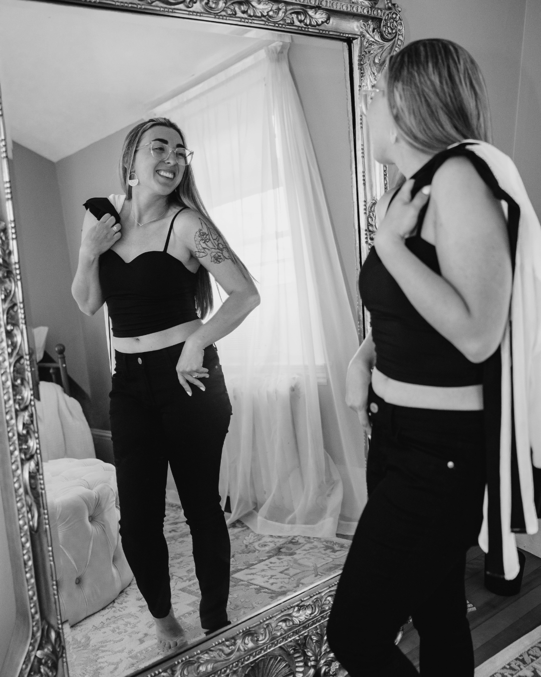 A woman with long hair, glasses, and a tattoo on her arm is smiling at herself in a large ornate mirror, wearing a black crop top and black pants. She is holding a shirt over her shoulder, and another woman, also in black, is looking at her with her back to the camera, holding her own shirt over her shoulder. The room has light-colored curtains, a patterned rug, and a tufted ottoman.