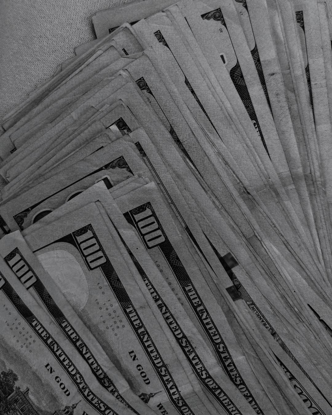 A black and white image of numerous US $100 bills fanned out and stacked.