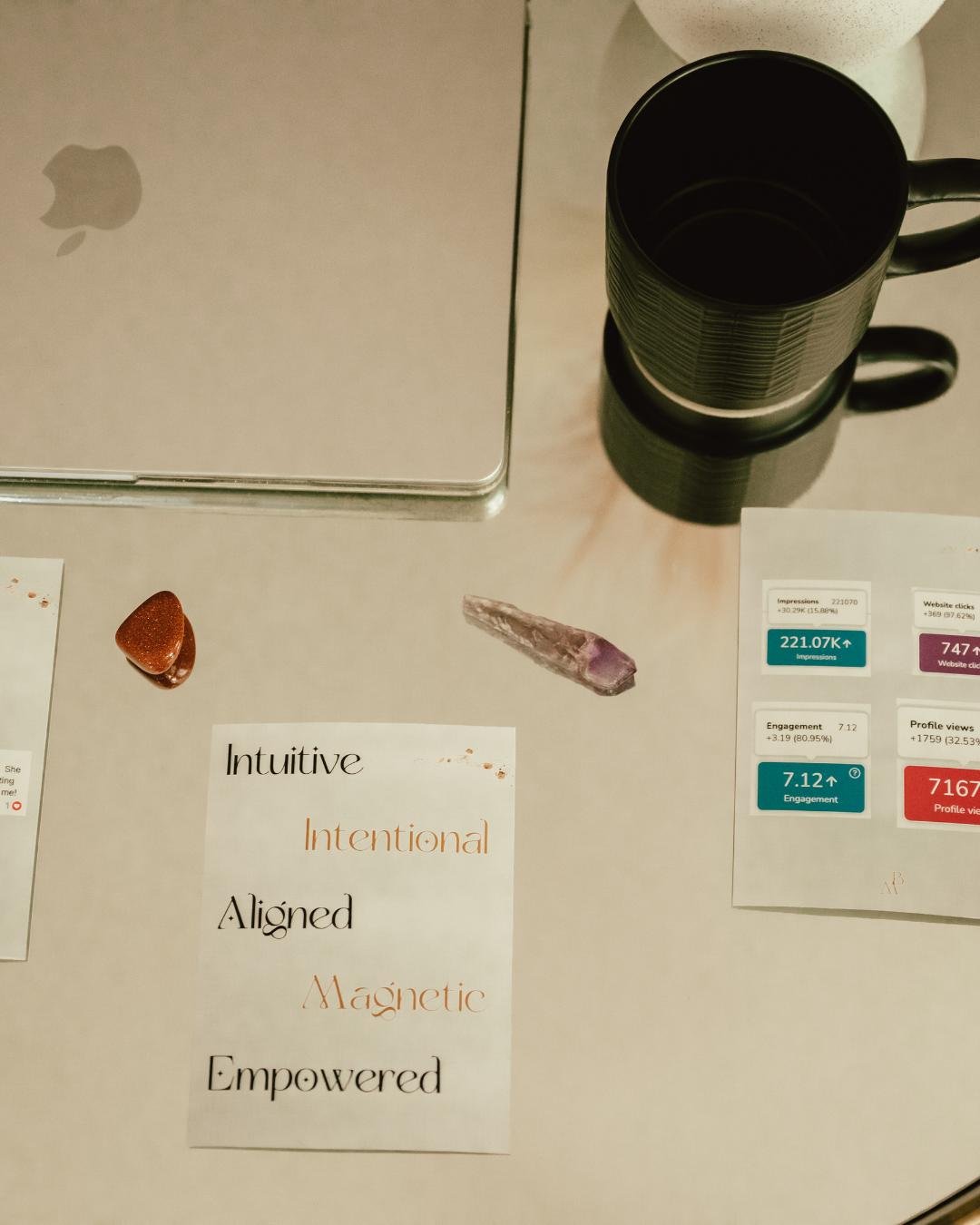 A desk with a closed silver Apple MacBook, a black mug, a small brown object, a purple crystal, and printed words that say 'Intuitive', 'Intentional', 'Aligned', 'Magnetic', and 'Empowered'