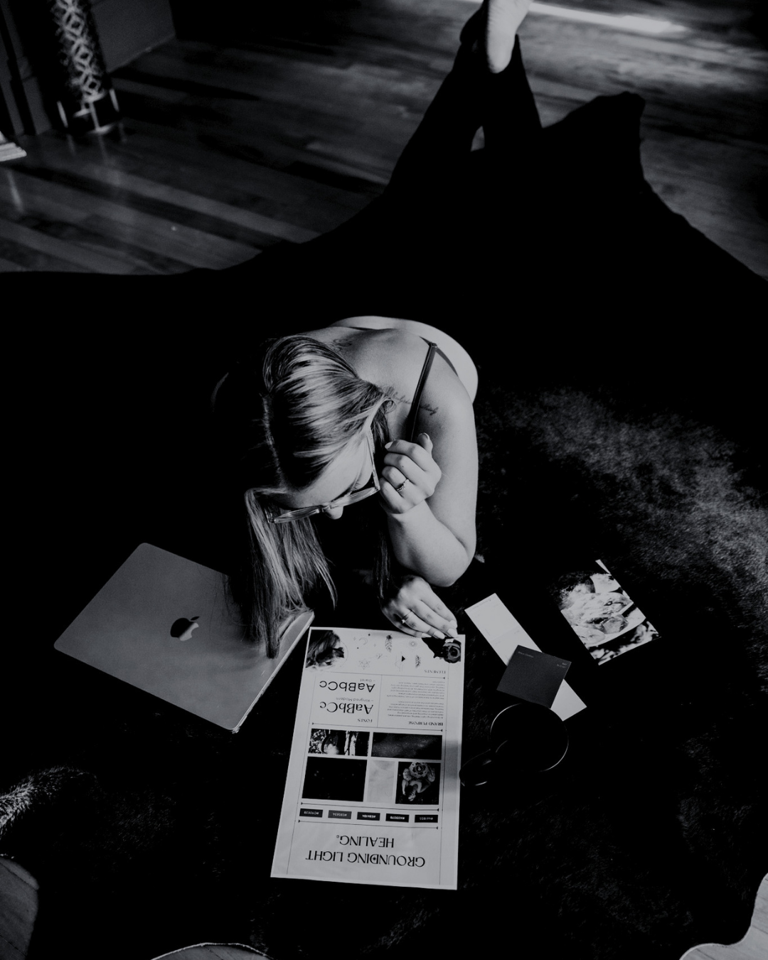 Black and white photo of a woman lying on the floor, reading a brochure titled 'Ground Light Healing' with a laptop, photographs, and a cup around her.