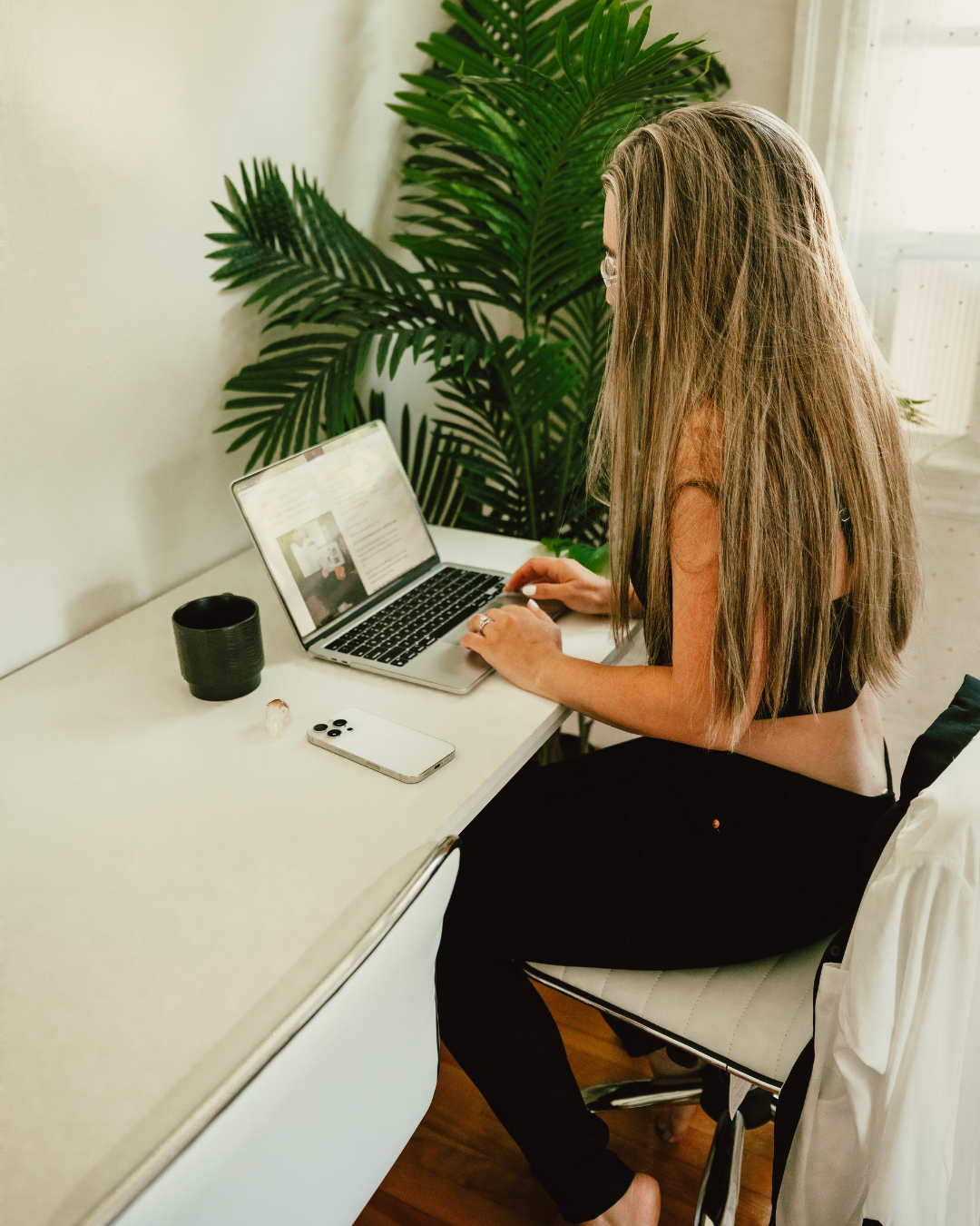 A woman with long, wavy hair and glasses working on a laptop at a white desk in a room with large green plants.