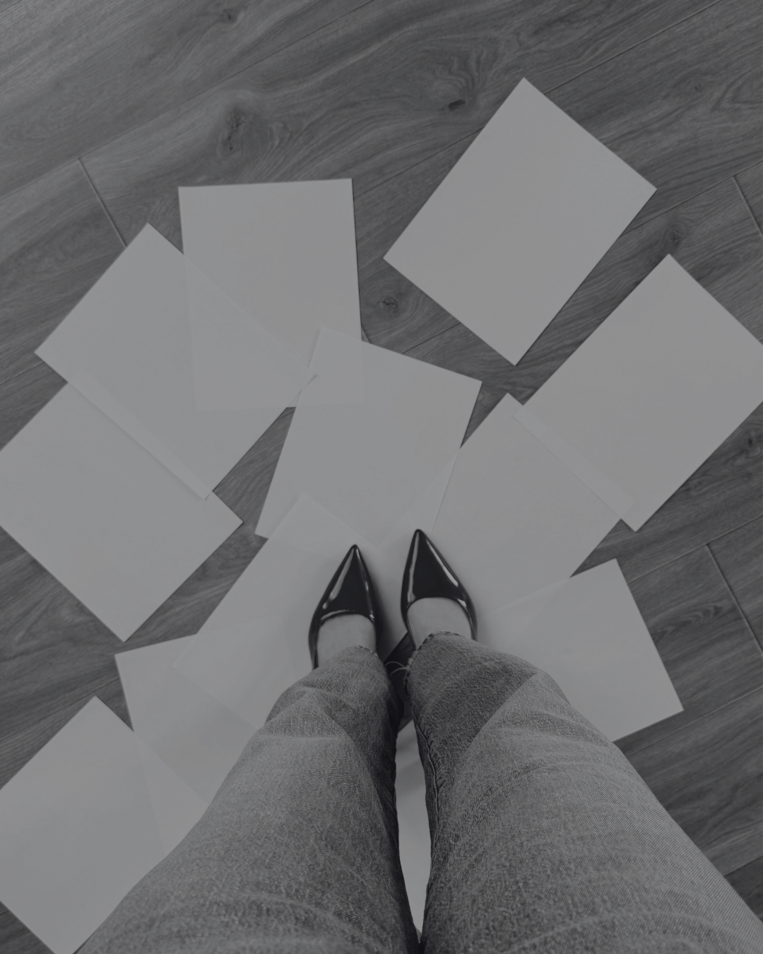 View from above of a person wearing black shiny high-heeled shoes and gray pants, standing on a wooden floor with scattered blank white sheets of paper around their feet.