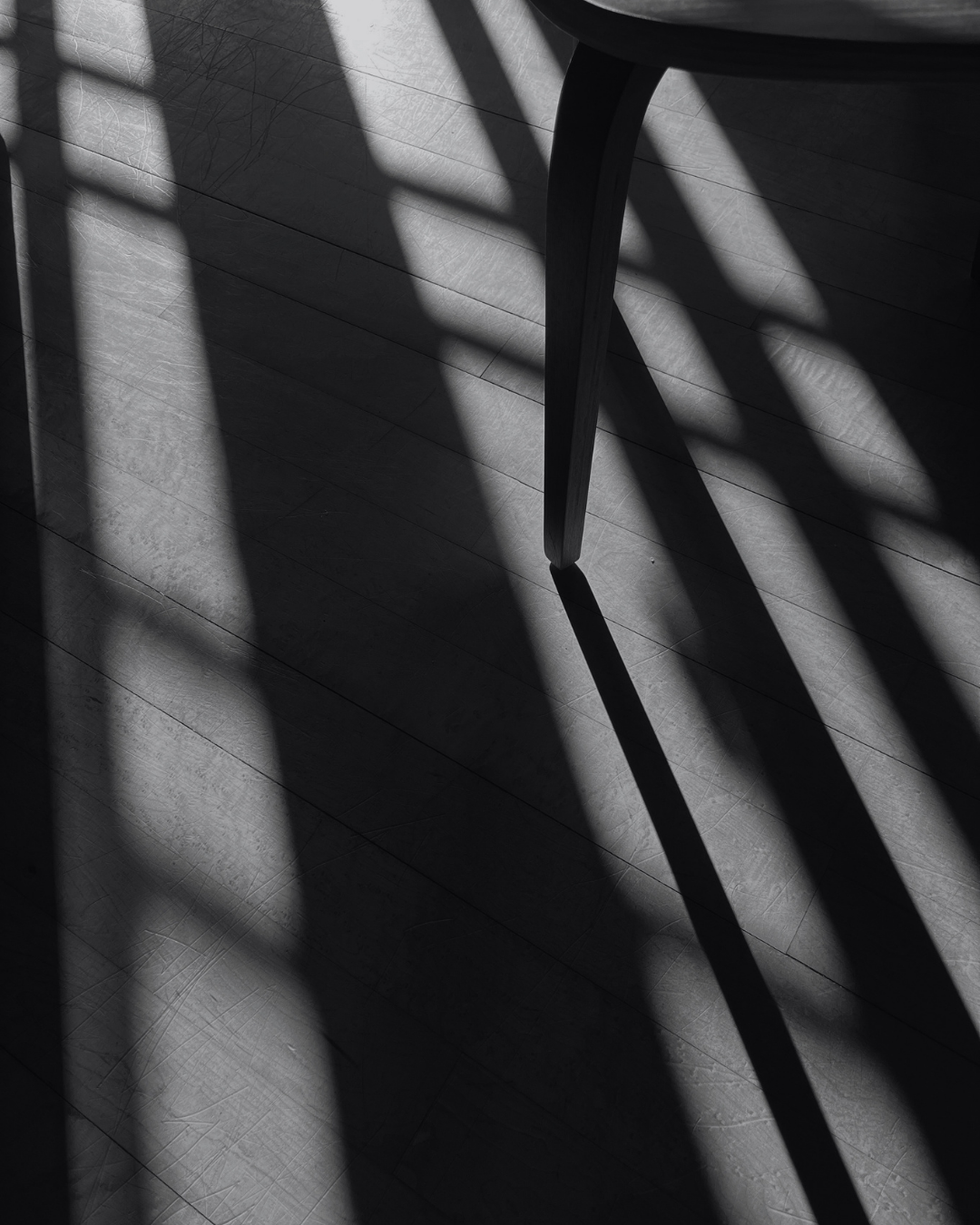 Black and white photo of sunlight casting shadows of a chair's slats onto a wooden floor, with a chair leg visible.