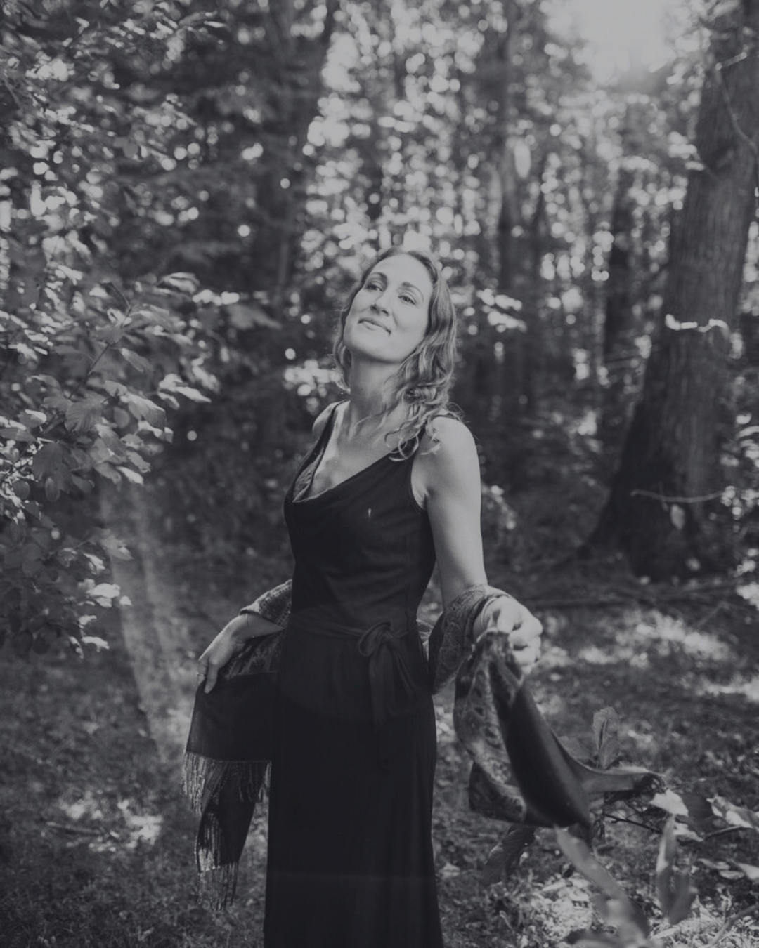 Black and white photo of a woman with wavy hair in a forest, wearing a sleeveless dress, and holding a scarf or shawl.