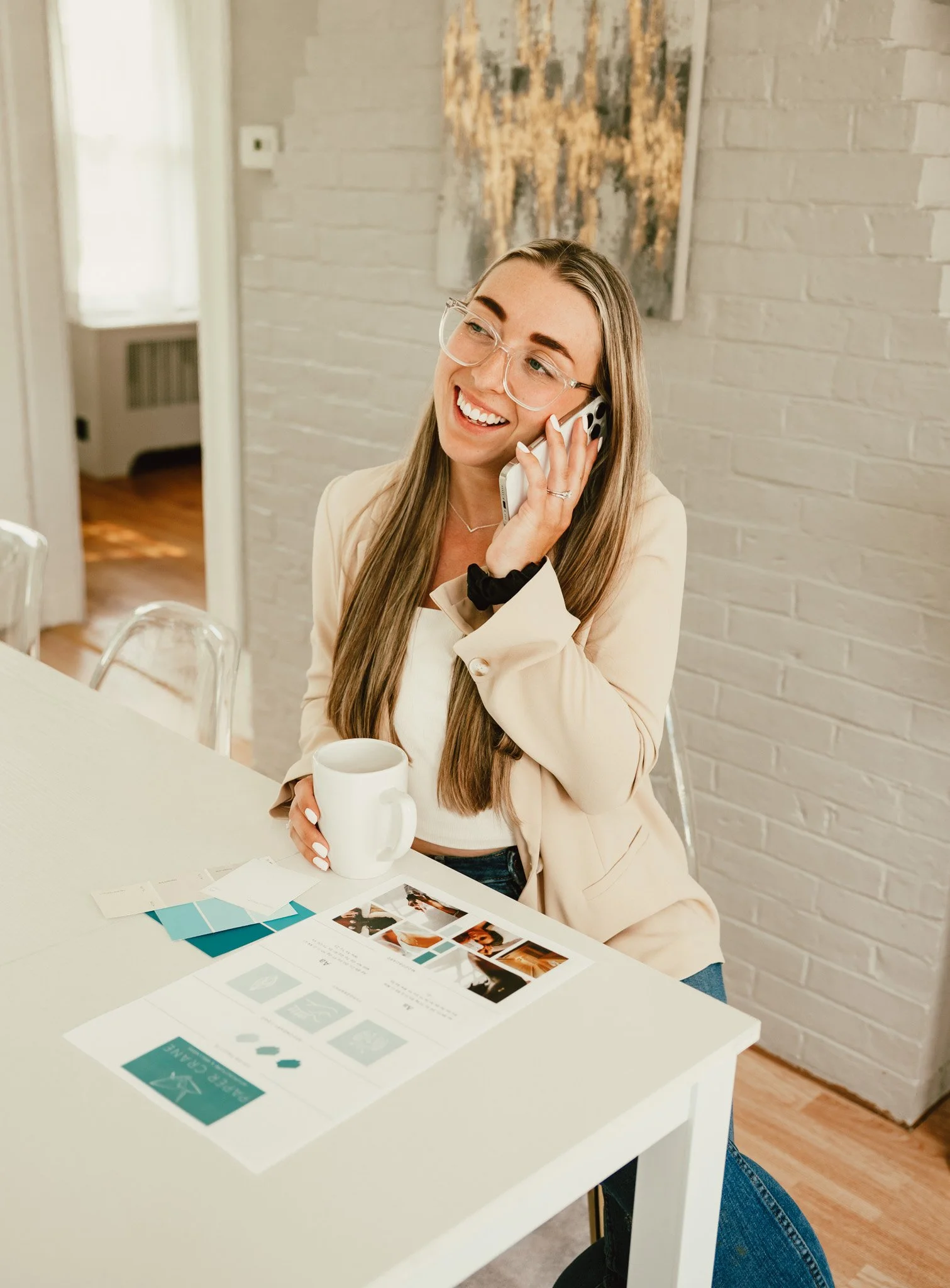 A smiling woman with long hair and glasses talking on a cellphone while sitting at a white table with brochures, color swatches, and a white mug, in a room with a white brick wall and a piece of artwork.