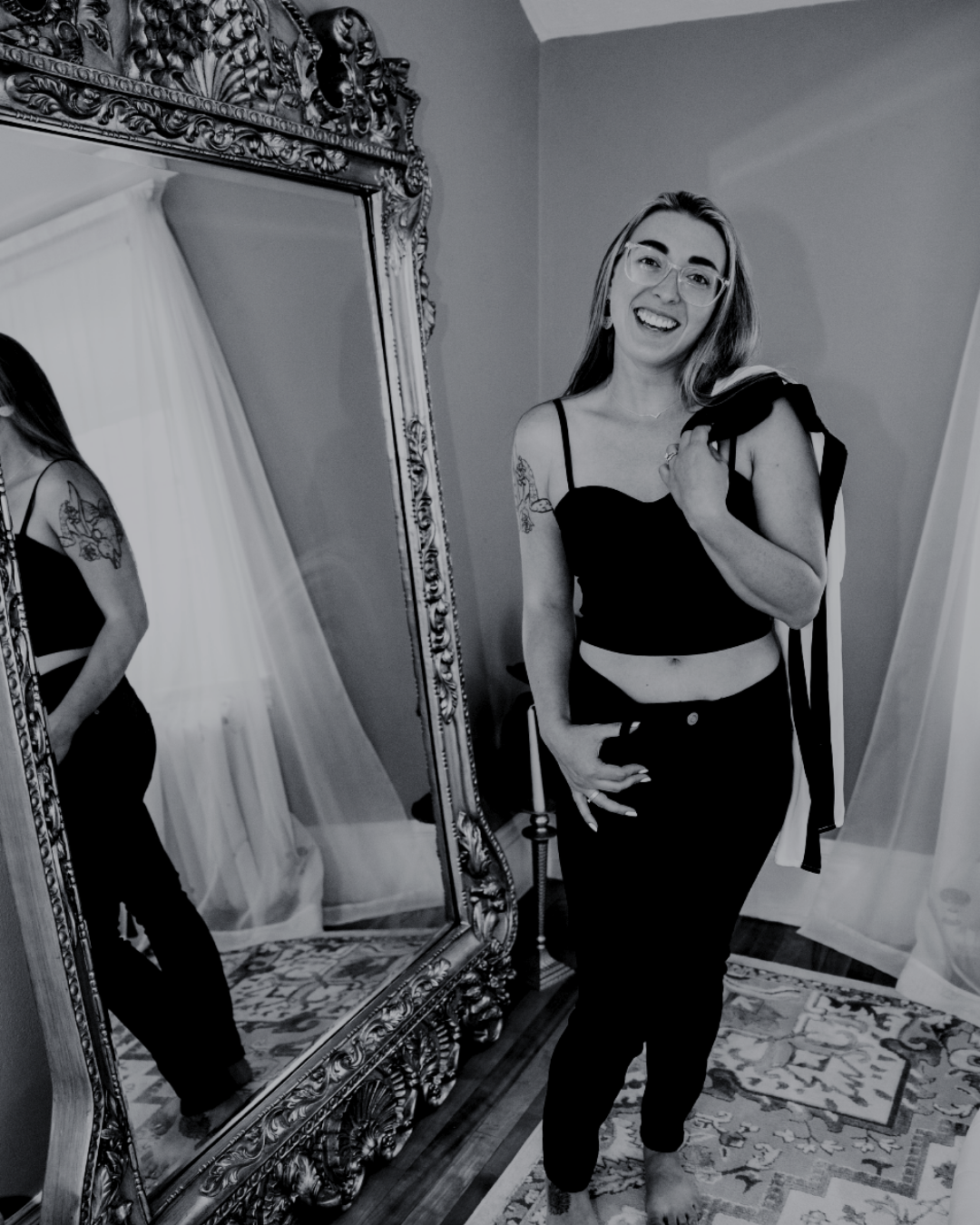 A woman with glasses and a tattoo on her arm is standing in a room, smiling at the camera, wearing a black crop top and black pants. She is holding a piece of clothing over her shoulder. The room has an ornate mirror, a patterned rug, and sheer curtains.