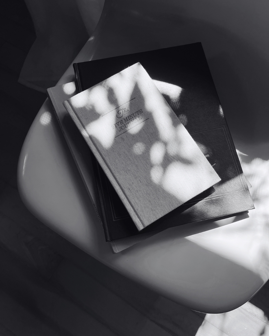 A stack of magazines and booklets on a small table, with sunlight filtering through a plant's shadow.