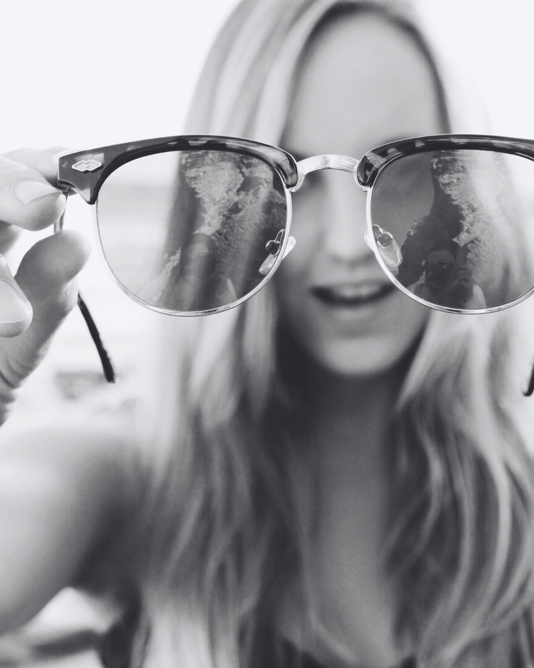 A woman holds sunglasses in front of her face, with the lenses reflecting the beach scene and her face partially visible behind the glasses.