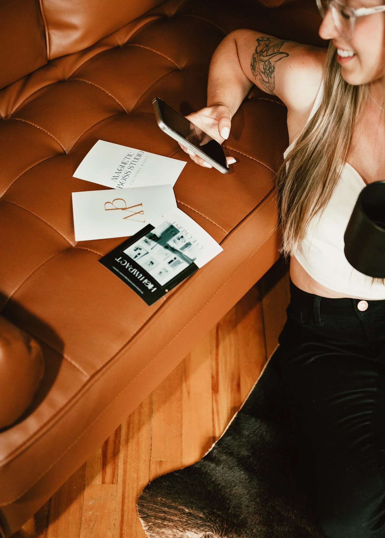 A smiling woman with glasses and long hair, lying on a brown leather couch, holding a smartphone. Several paper pamphlets are on the couch, with visible text including 'MASCULINE BOSS STUDY' and 'JAD'. The woman is wearing a white top and black pants, with a tattoo on her arm.