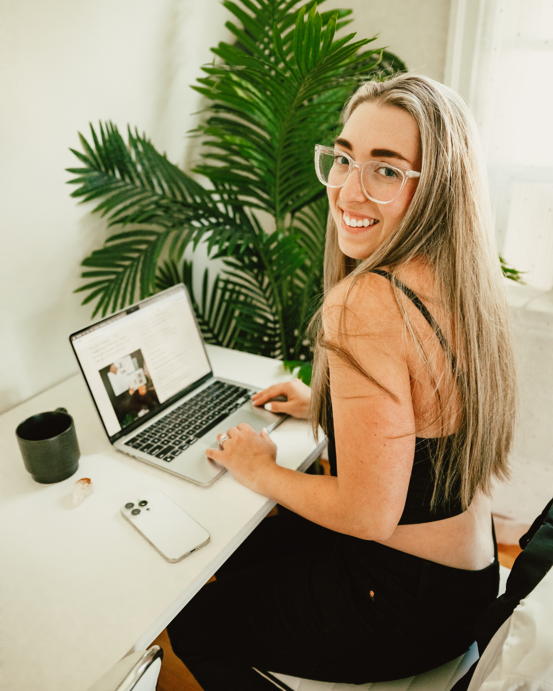 A young woman with long brown hair and glasses smiling at the camera while working on a laptop at a white desk. There is a large green potted plant in the background.