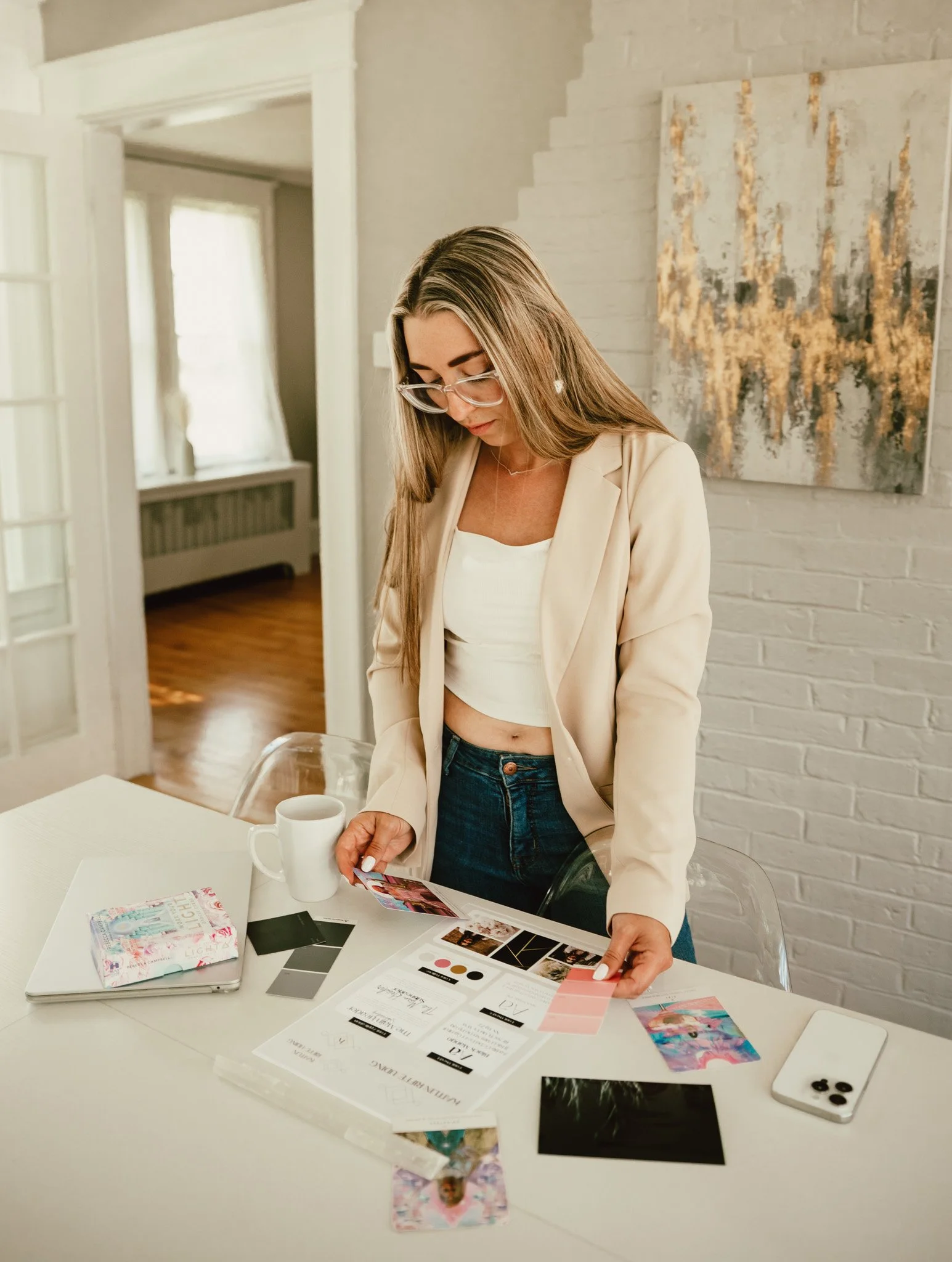 A woman with long blonde hair and glasses looking at a color palette and printed material on a white table with a white coffee mug and smartphone. The room has white walls, large windows, and a modern abstract painting on the wall.