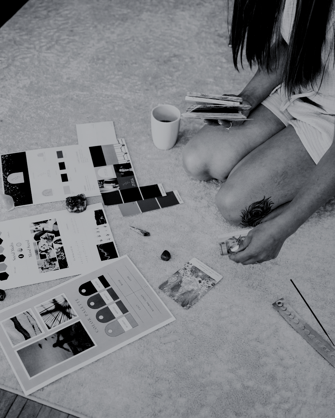 A person is sitting on a carpeted floor, examining magazines and pamphlets, with a coffee mug nearby, and various artistic supplies and stones spread around.