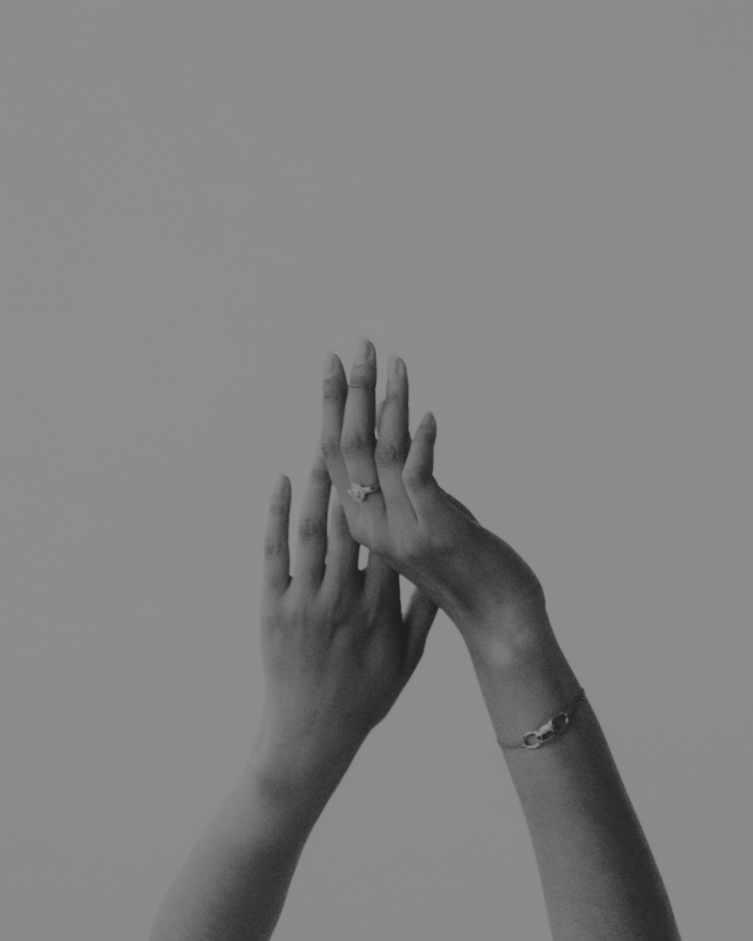 Two hands overlapping, displaying a ring on the ring finger, with bracelets on the wrists, against a plain background.